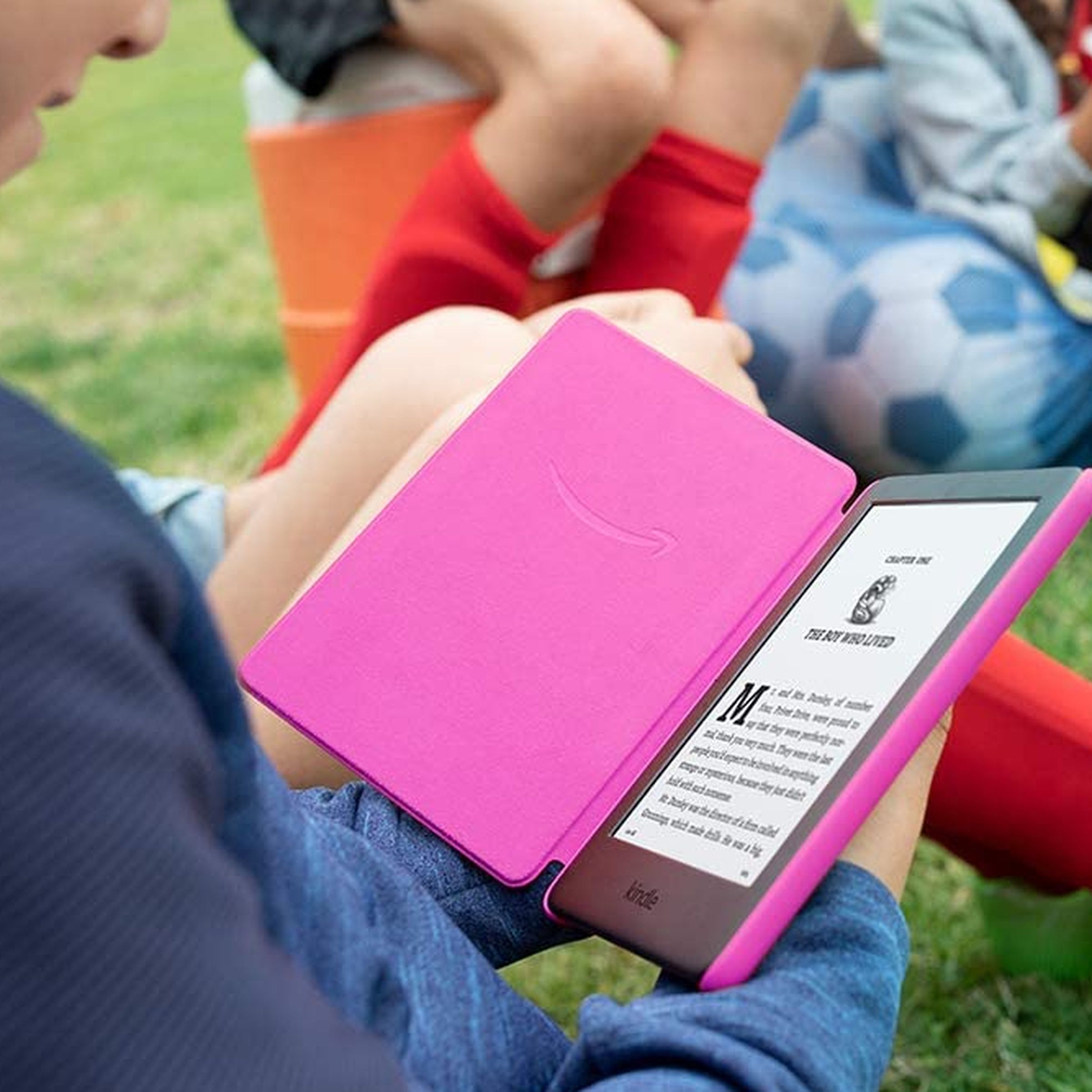 طفل يحمل جهاز Kindle ورديًا ويقرأه أثناء جلوسه على العشب.