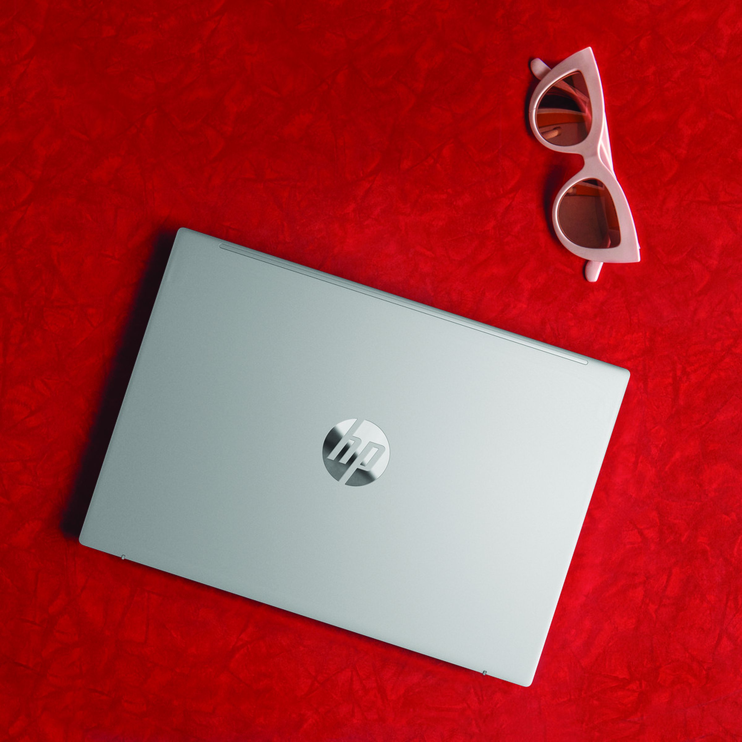 The HP Pavilion Aero 13 closed on a red background. To the right is a pair of pink sunglasses.