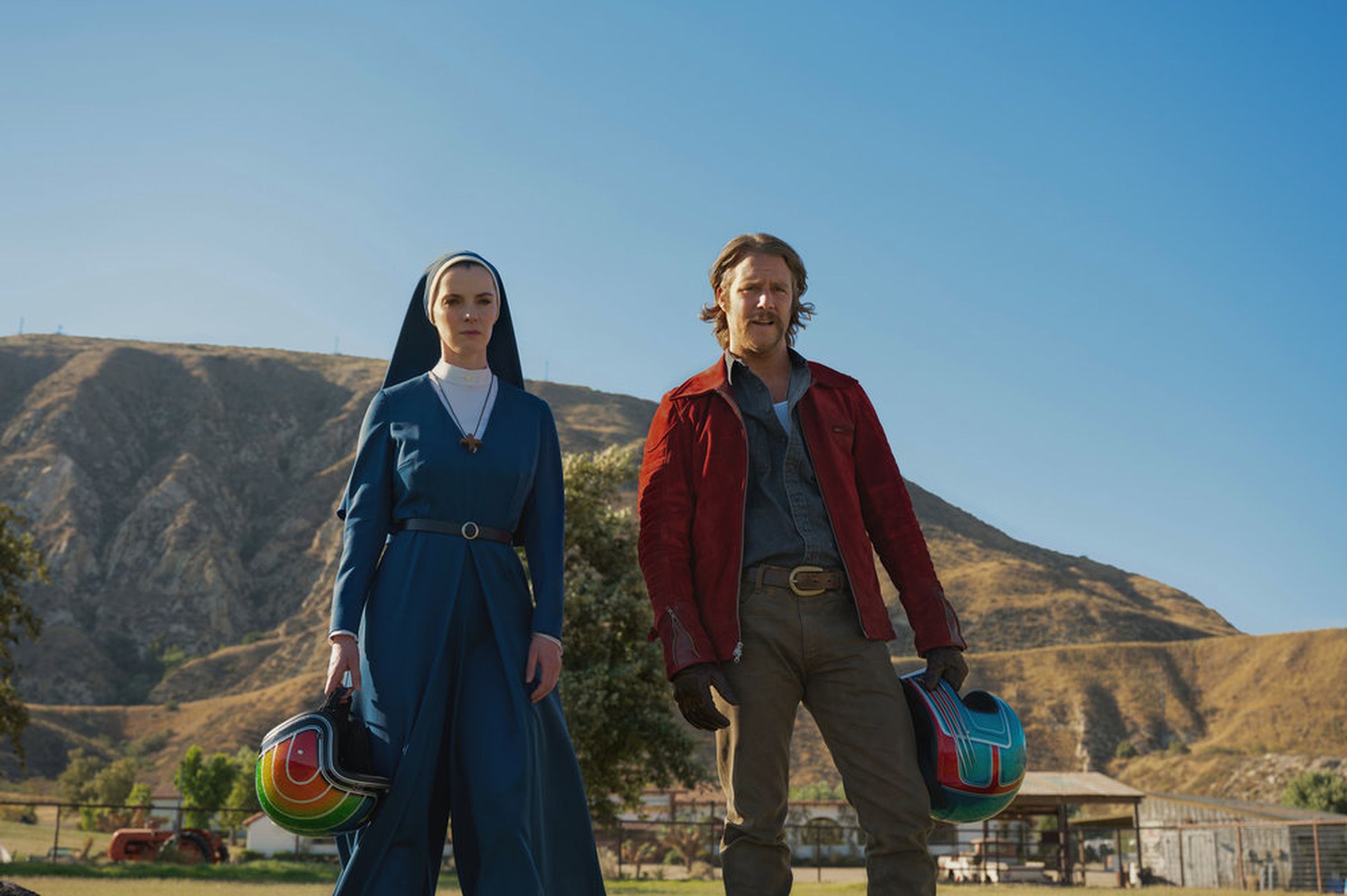 A white woman dressed as a nun and a white man with blond hair and a mustache, stare at something in concern. They both hold colorful motorcycle helmets.