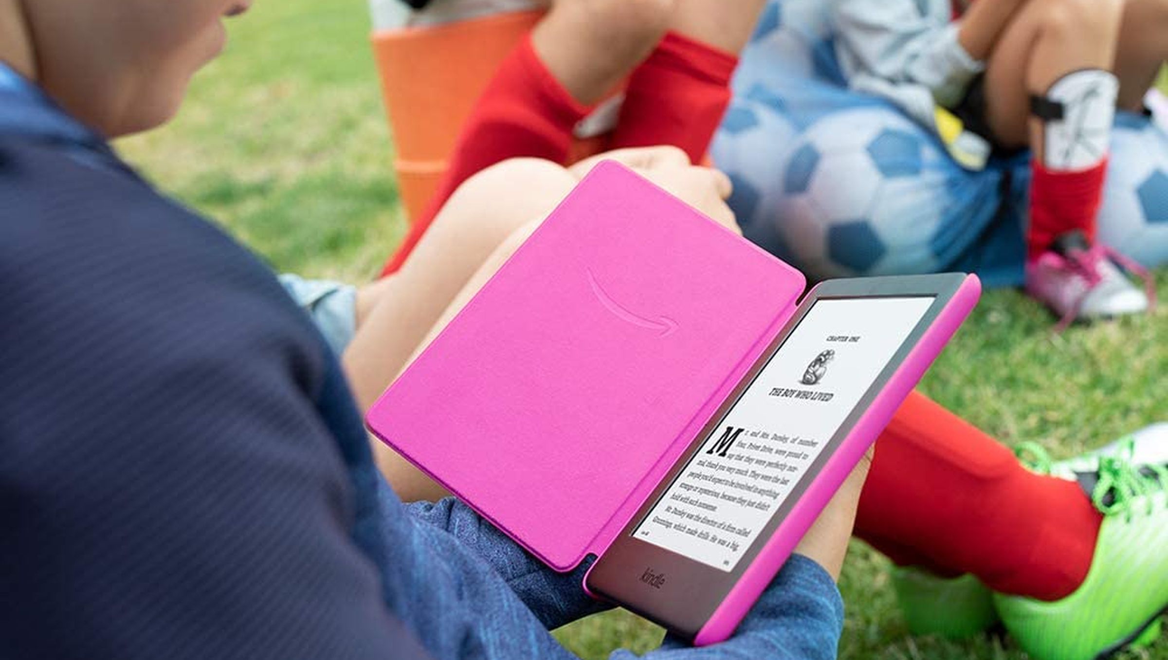 A child holding and reading a pink Kindle while sitting on grass.