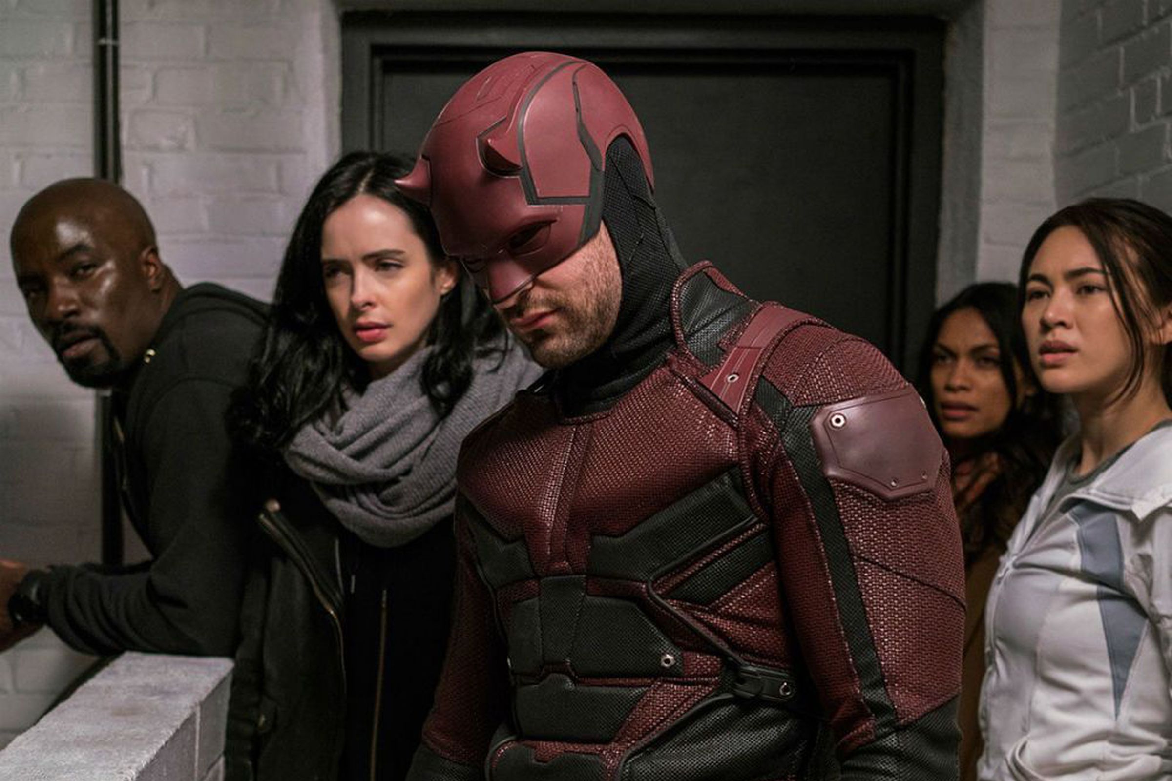 A man in an armored, red and black superhero suit including a helmet adorned in small devil horns. The man is flanked to his right by a bald man in a gray hoodie, a brunette woman wearing a leather jacket and a chunky scarf. To the man in red’s left by a woman in a black top, and another woman in a white jacket.