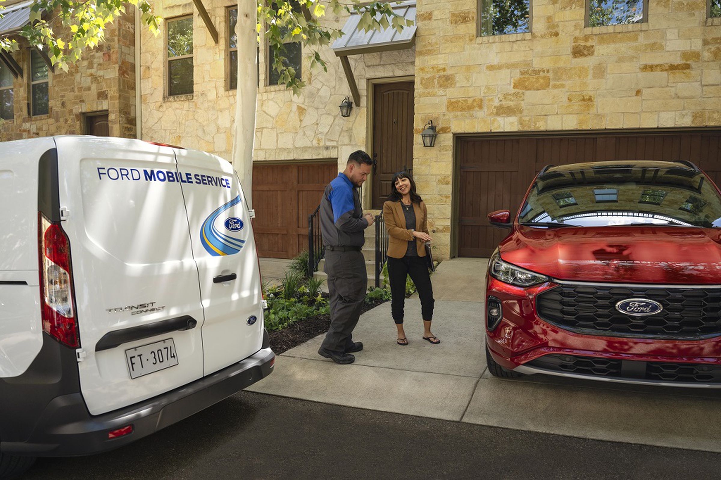 a white ford mobile service truck is parked on the street in front of a brick and stone home with a red ford vehicle in the driveway. A woman wearing a burnt Sienna blazer is gesturing towards her car for a man in automobile service uniform. She’s holding a laptop, probably because she works from home.