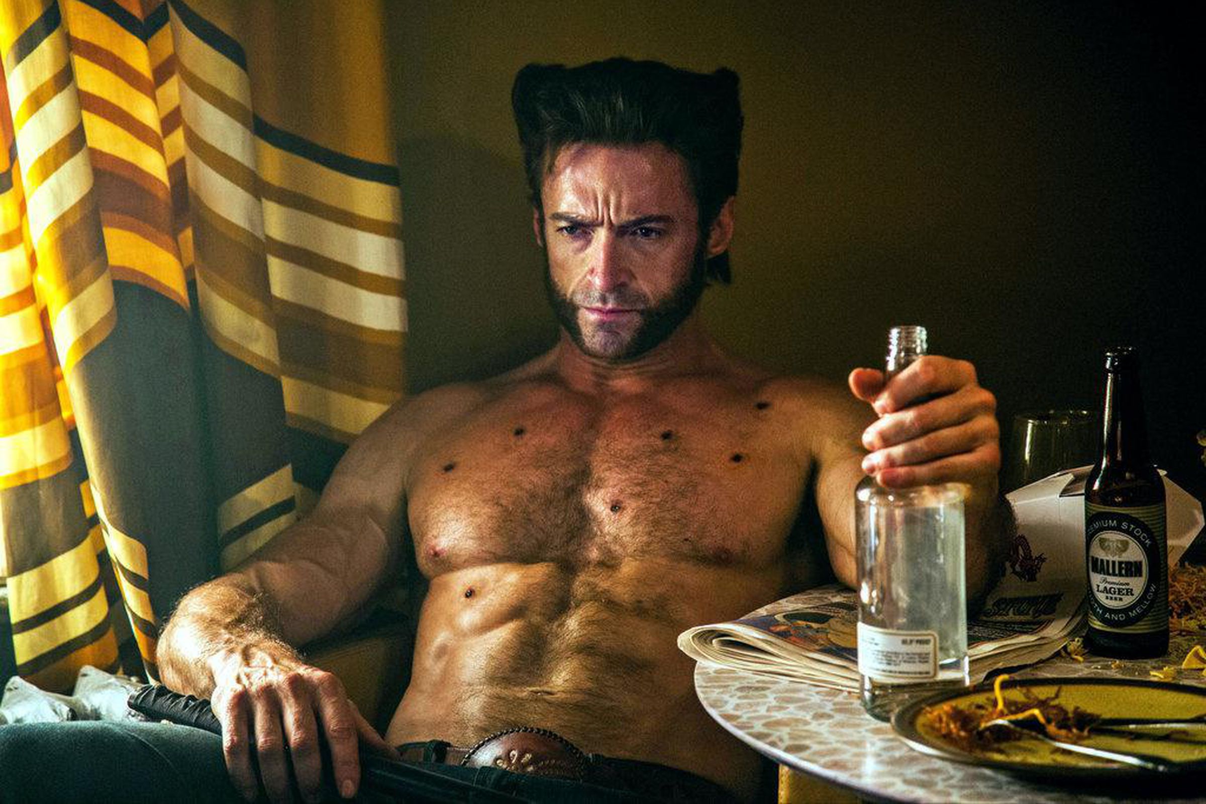 A shirtless man riddled with bullet wounds that are healing very quickly as he sits in a dingy hotel room drinking hard liquor.