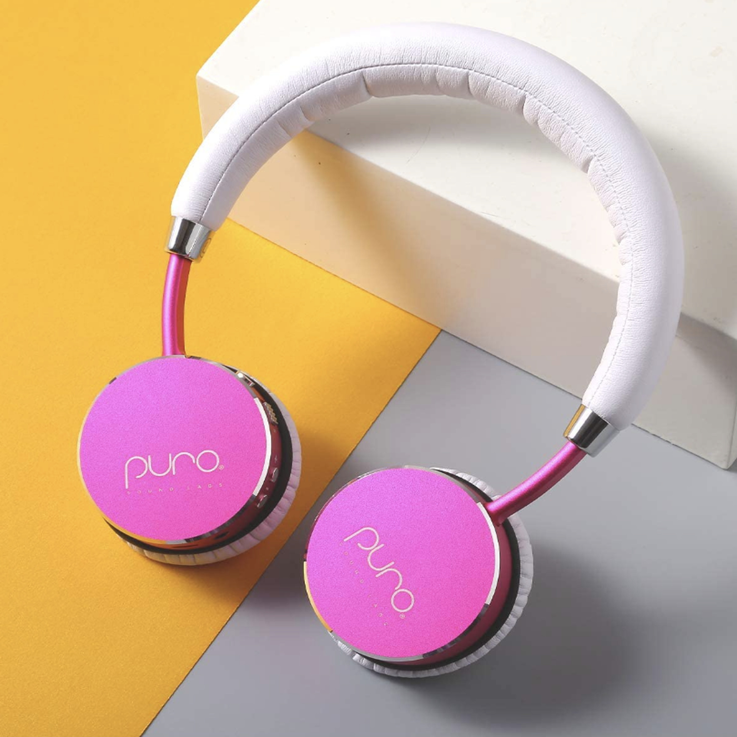 Headphones with white headband and pink over-the-ear cushions on a yellow, gray and white background.
