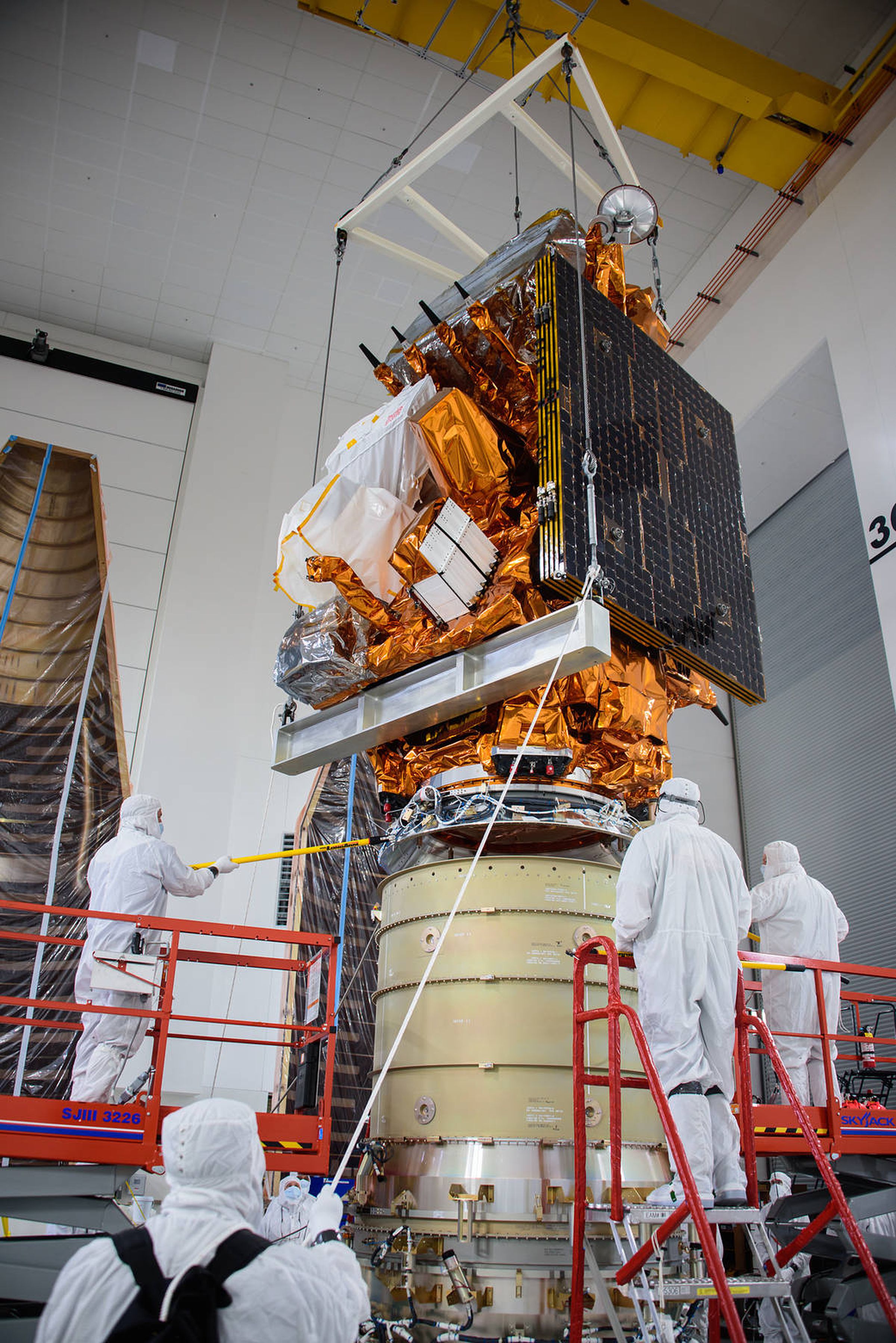 Vier Personen in weißen Anzügen stehen in einem Reinraum um einen Satelliten herum