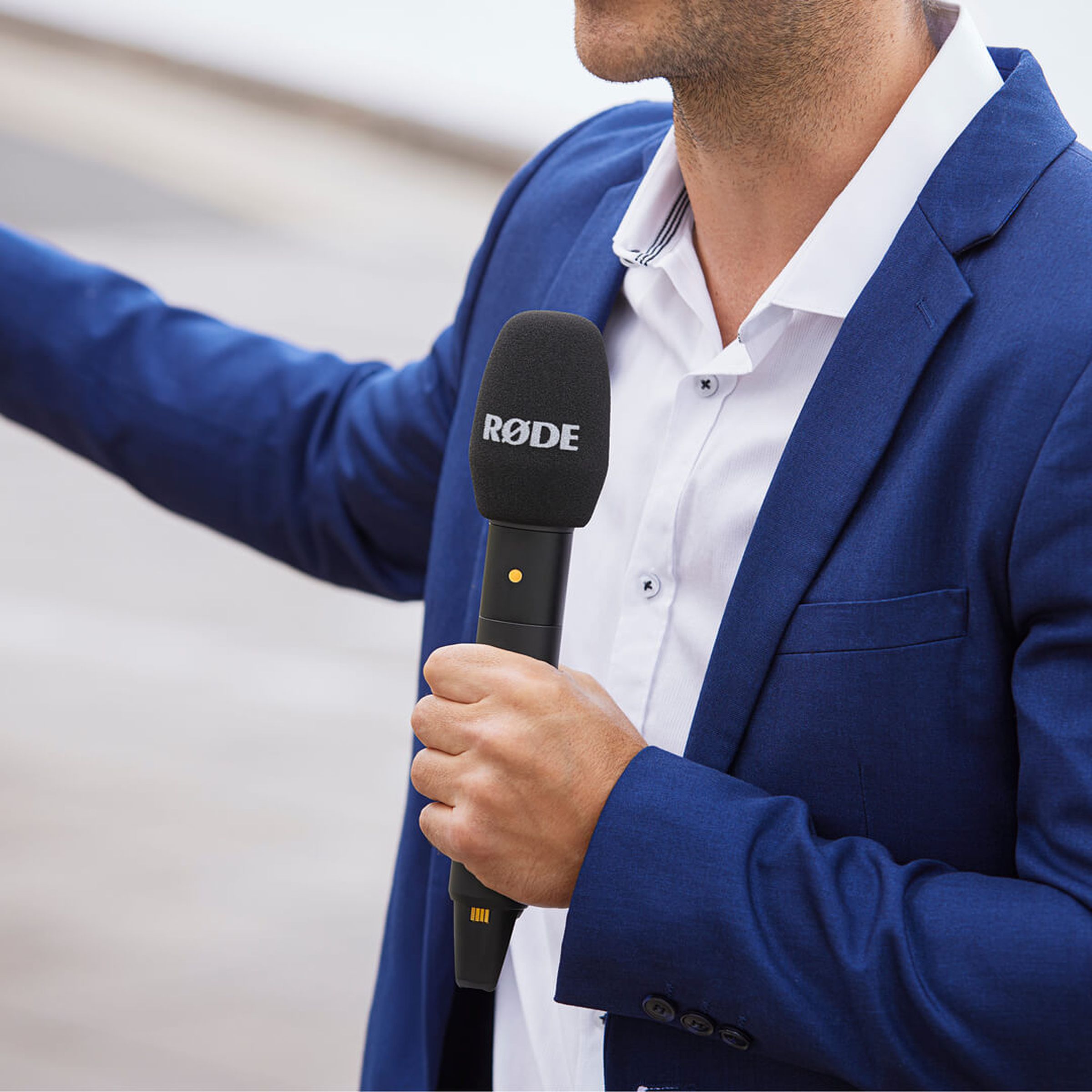 A person in a blue blazer holding a big stick microphone outside.