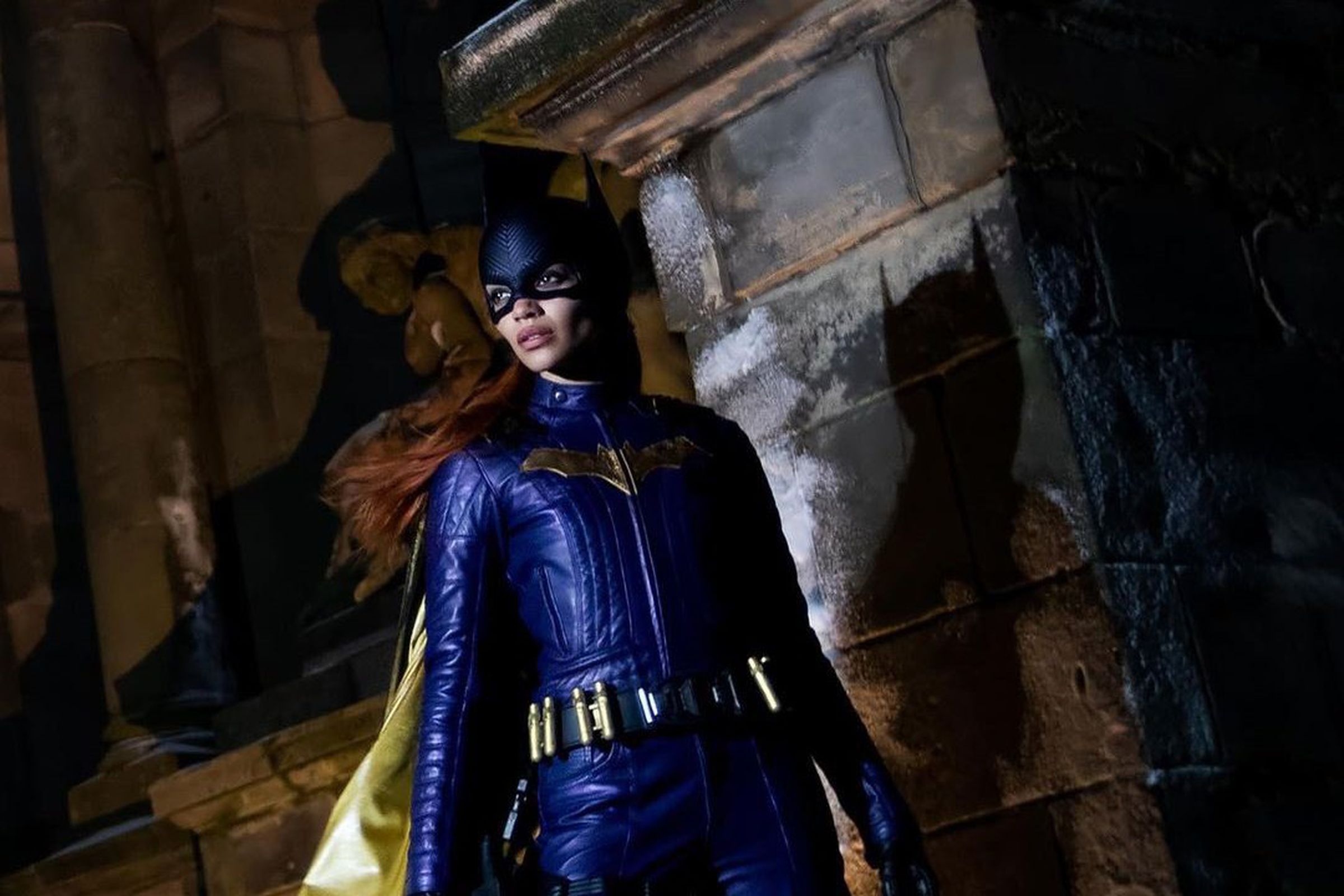 Leslie Grace is wearing a red wig and a purple Batgirl costume and mask. She appears to be standing on a rooftop.