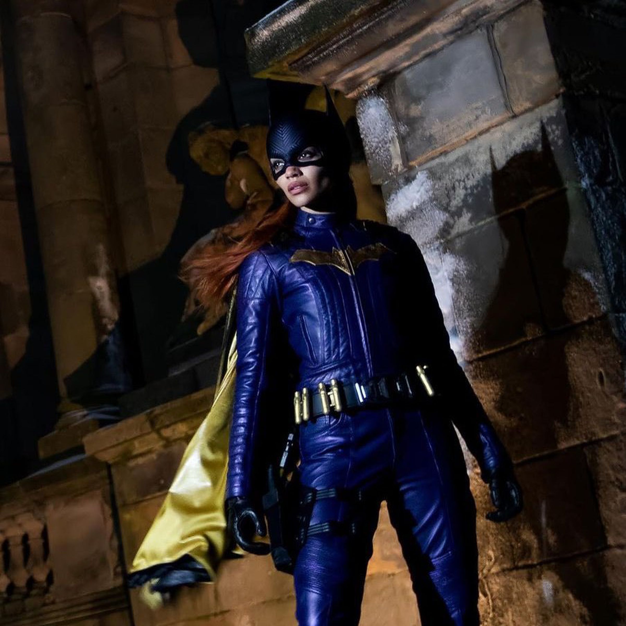 Leslie Grace is wearing a red wig and a purple Batgirl costume and mask. She appears to be standing on a rooftop.