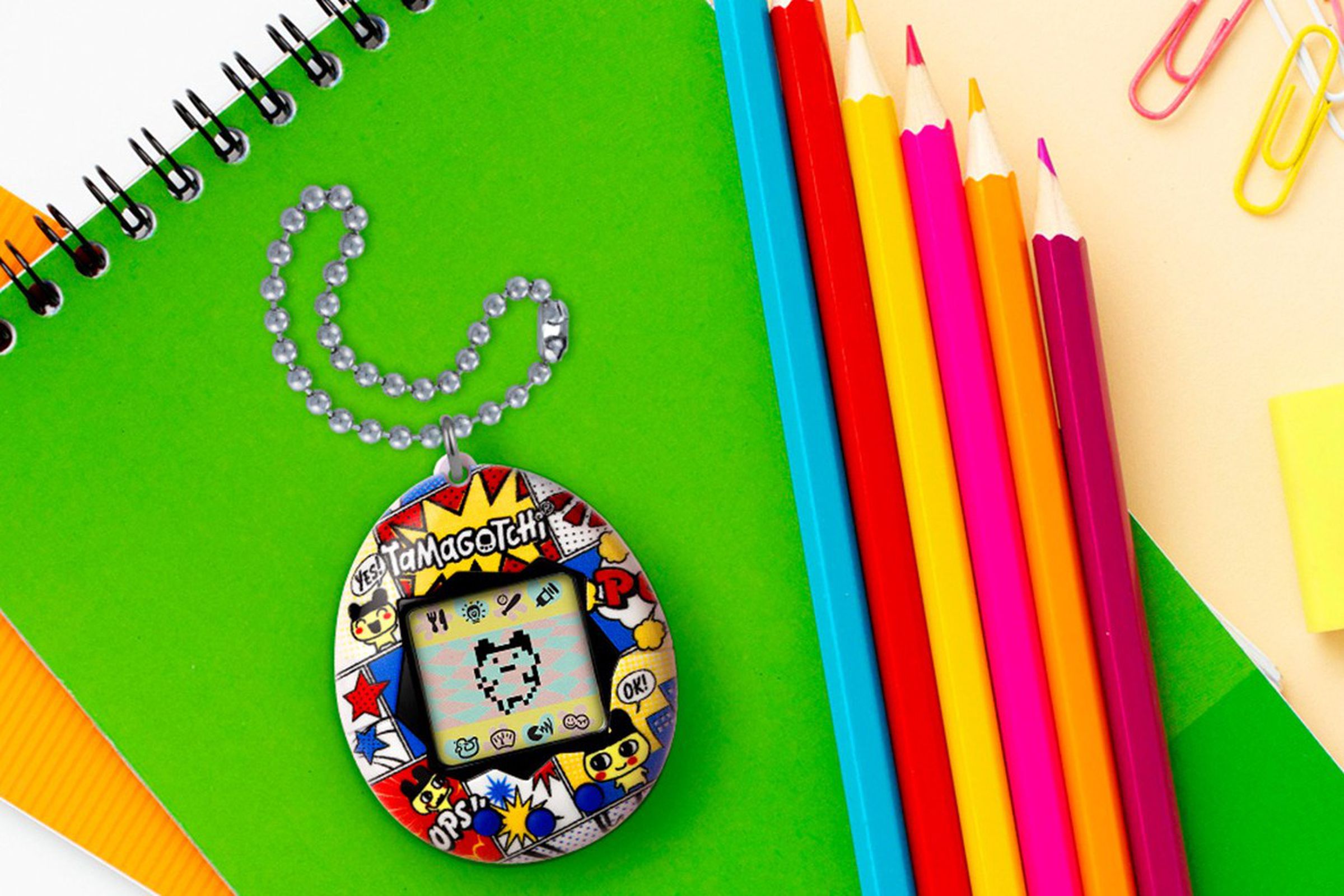 A bright and colorful Tamagotchi resting on a green notebook near colored pencils.