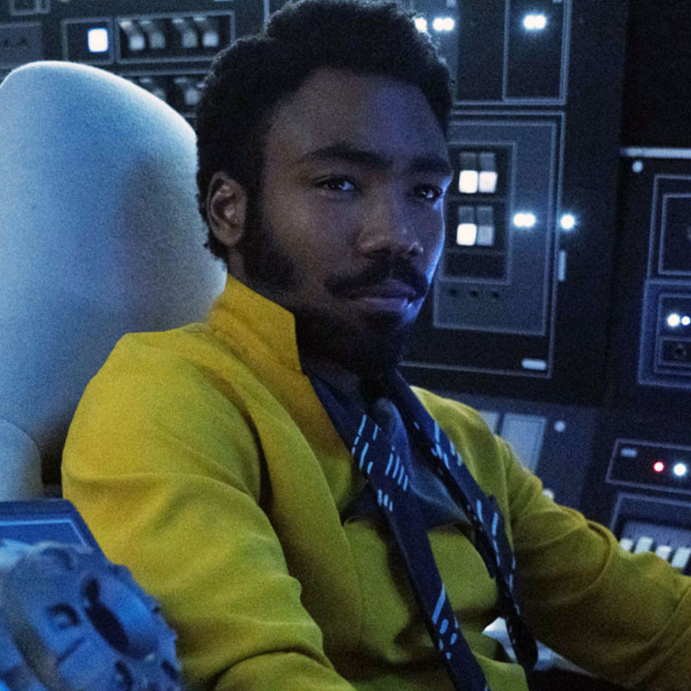 A stylish man in a yellow shirt sitting in the cockpit of a spaceship surrounded by an array of controls.