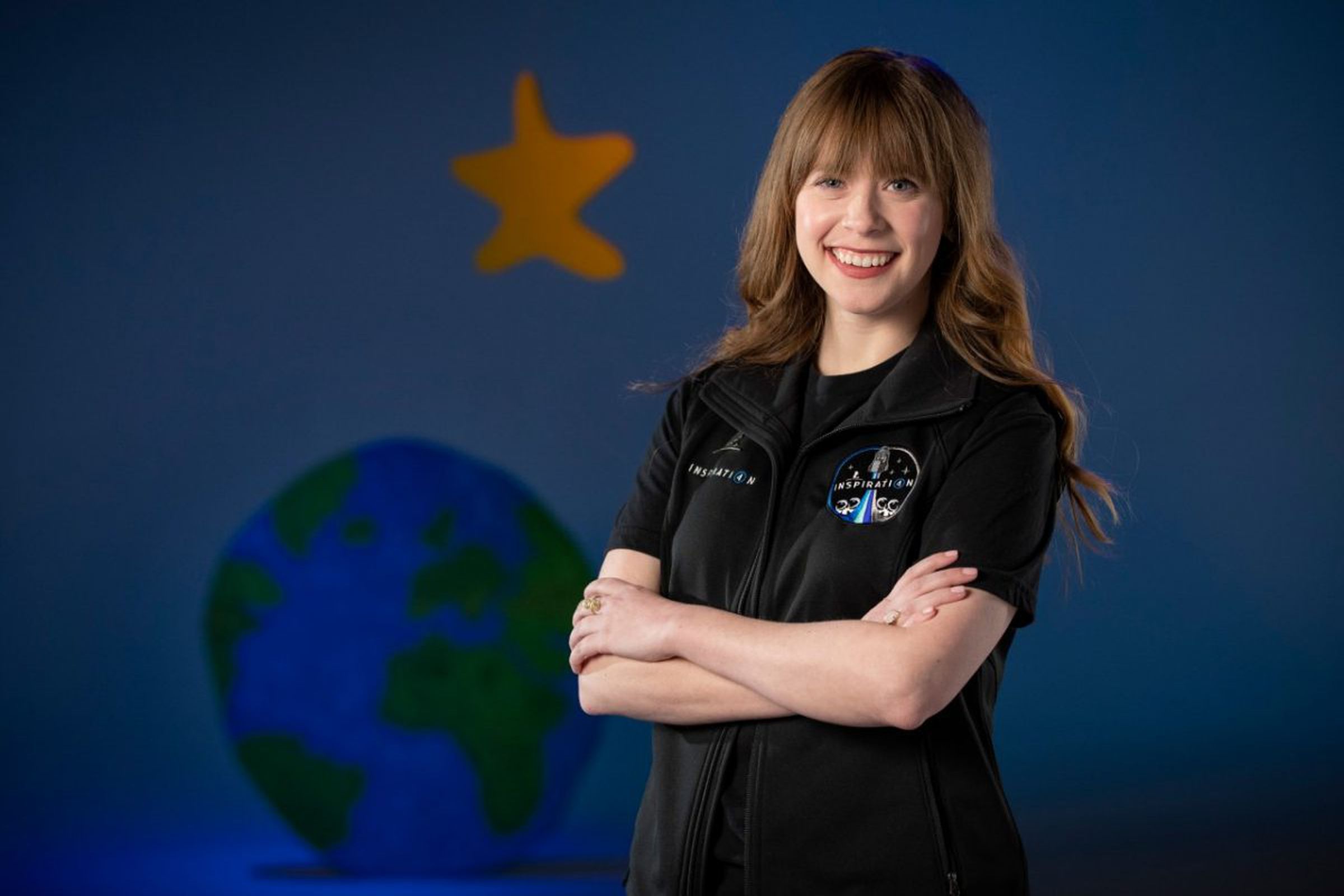 A white woman with long, light brown hair smiling and standing with her arms crossed in front of a wall painted with planet earth and a star.