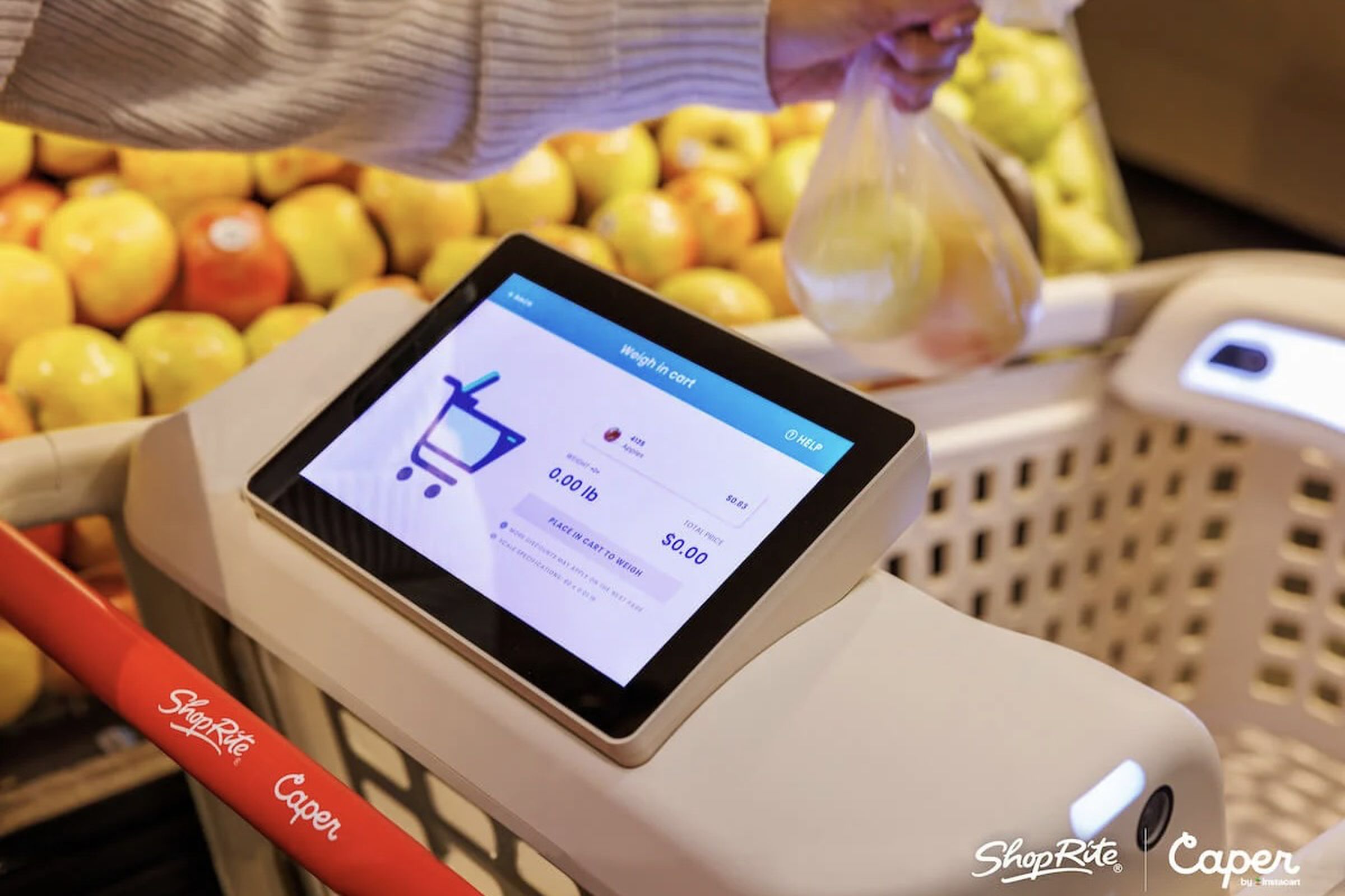 grocery cart with a tablet build onto it as a person lowers a bag of produce that will be weighed automatically by the cart