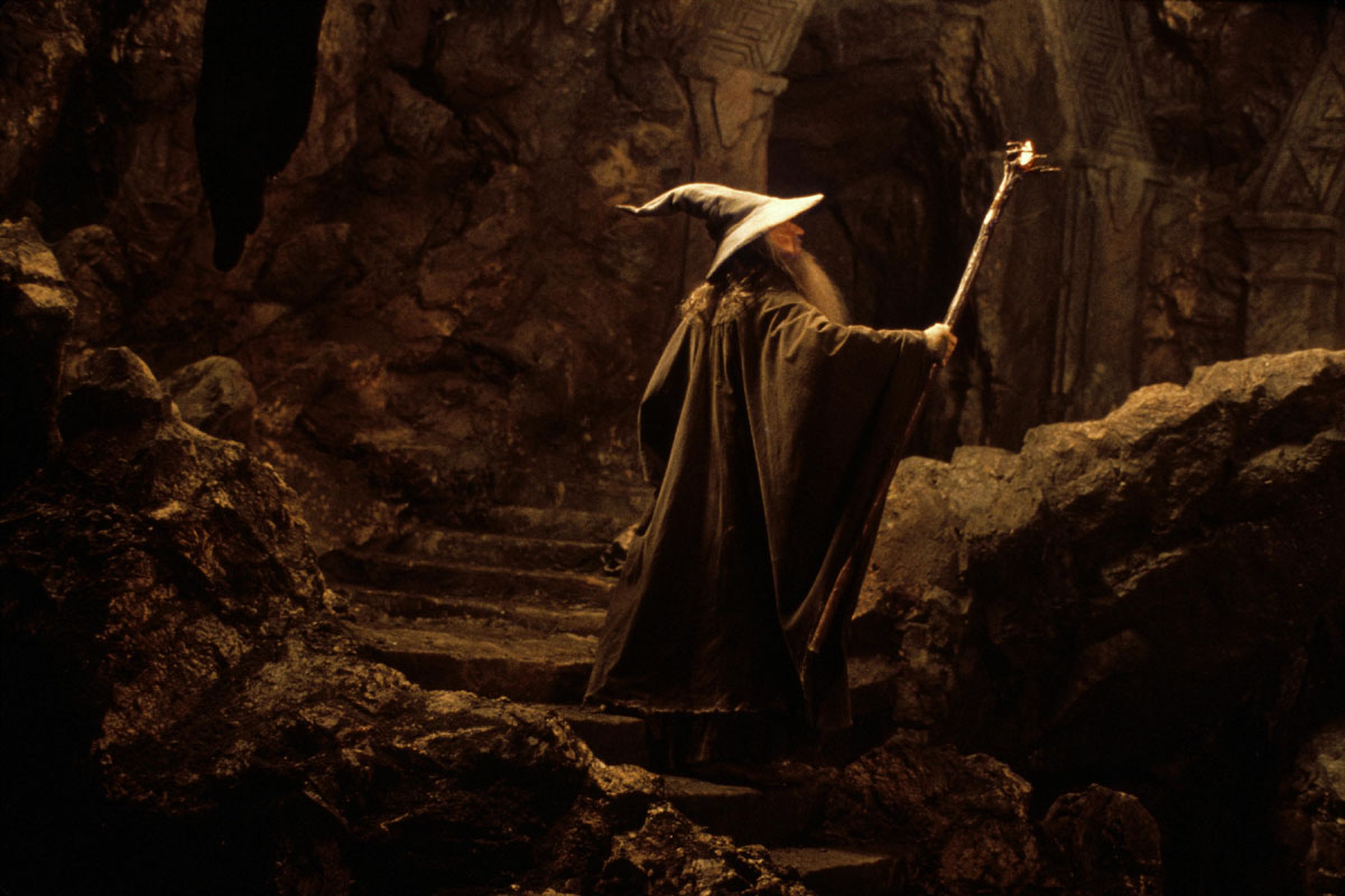 Gandalf the Grey garbed in a gray robe holding up his stave in a dimly lit cave