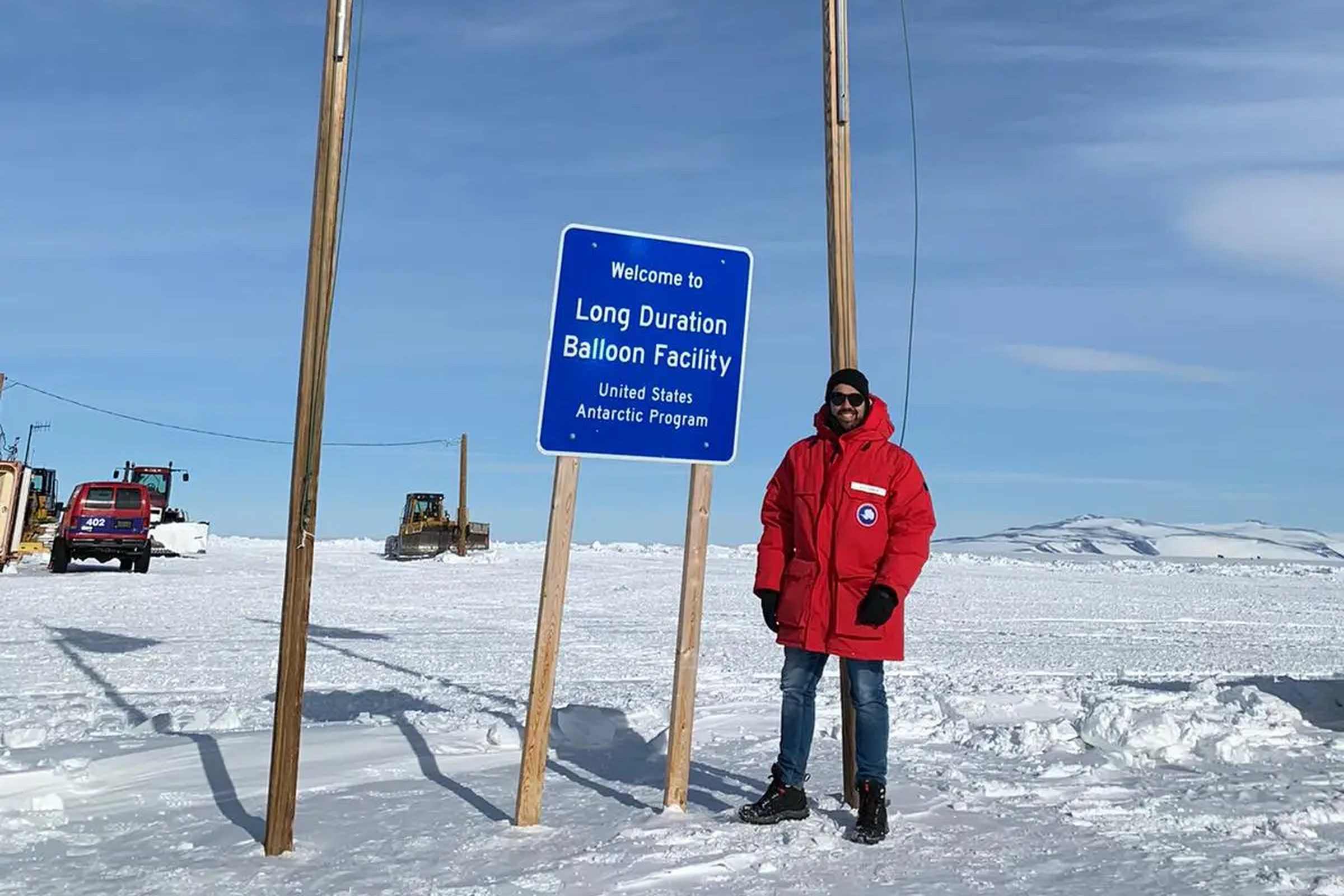 A picture of a person standing on snow next to a sign reading “Welcome to Long Duration Balloon Facility.” Smaller letters beneath say “United States Antarctic Program.”