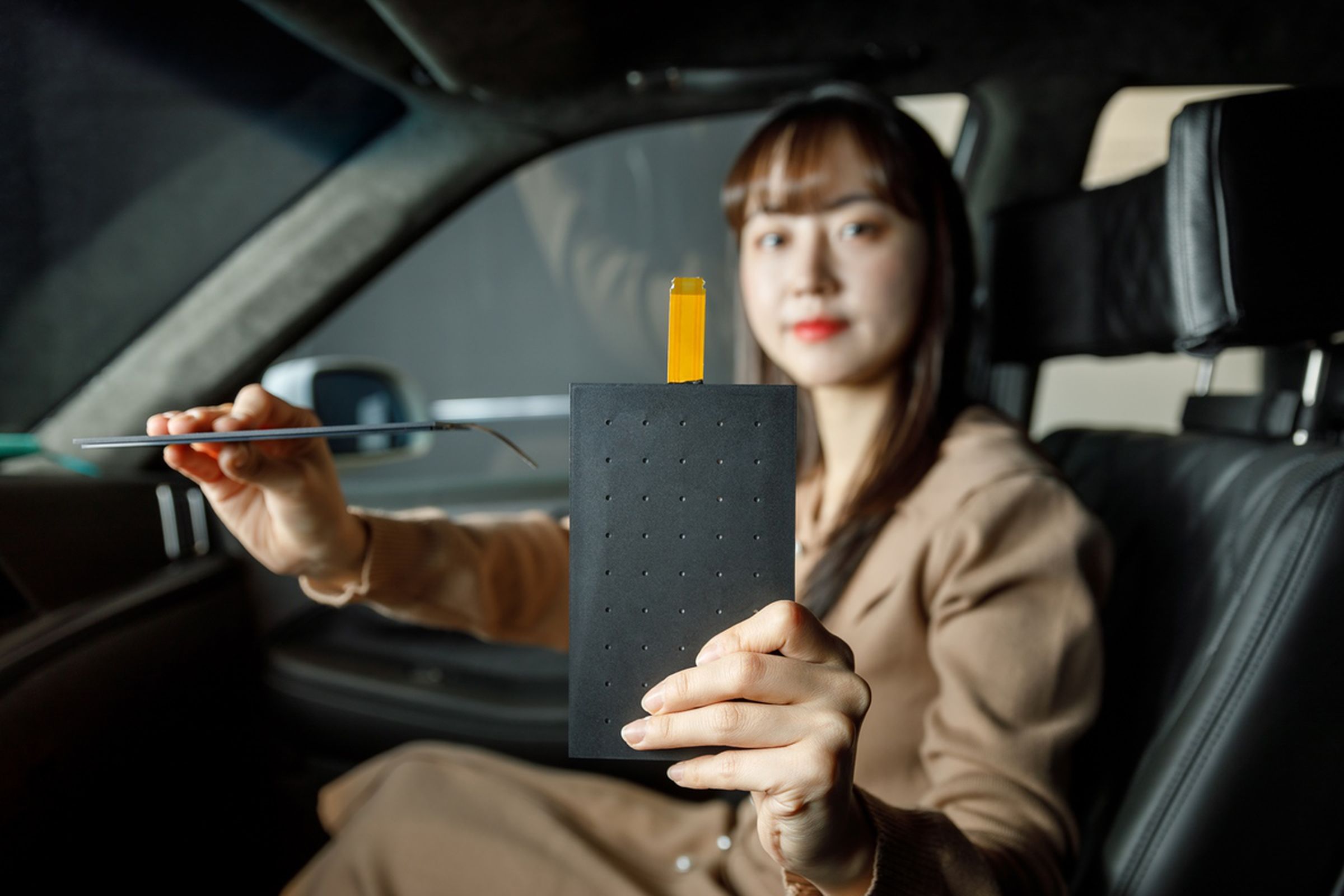 A woman in a car, holding LG Display’s Thin Actuator Sound Solution panel. The panel is a slim, black slab of plastic-looking material.