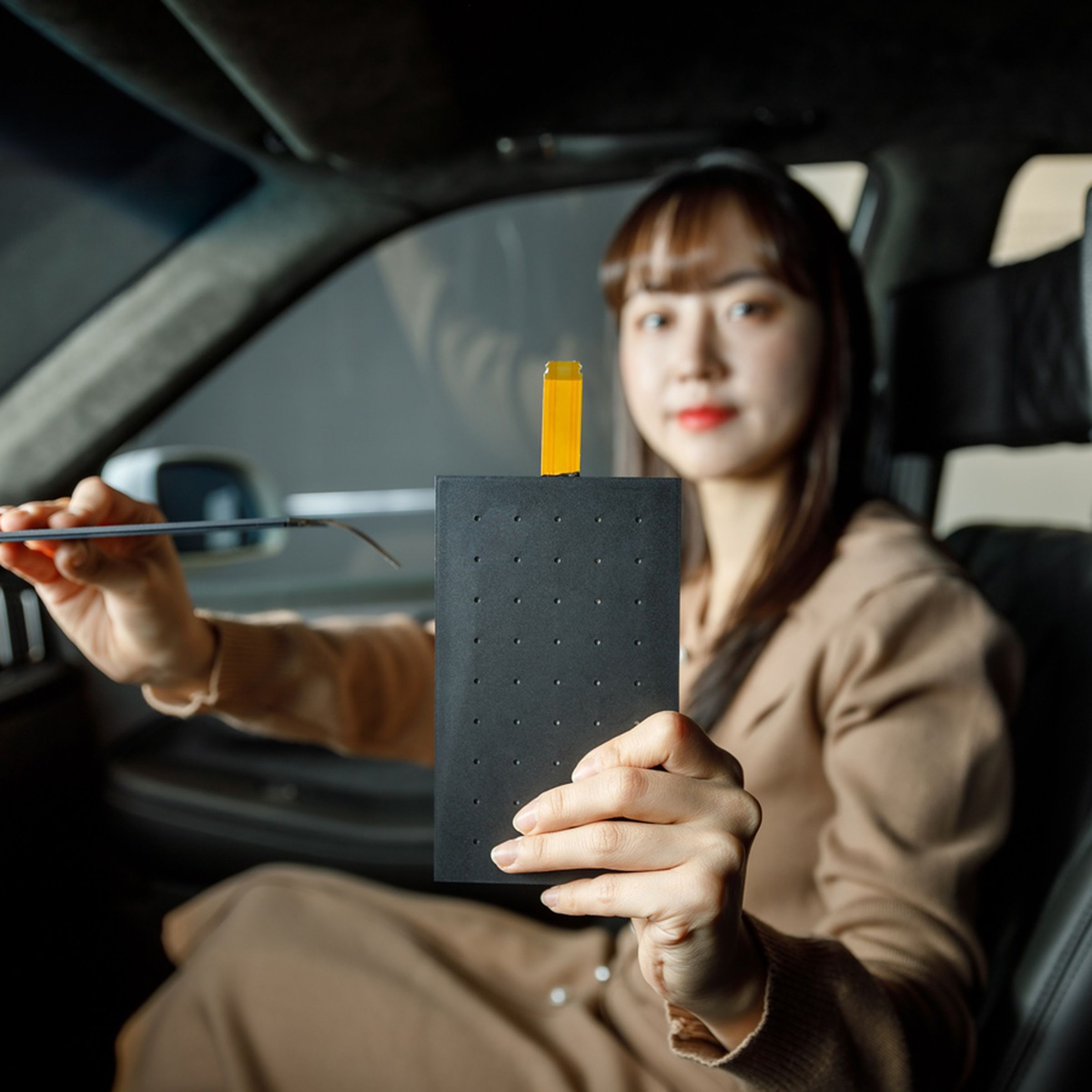 A woman in a car, holding LG Display’s Thin Actuator Sound Solution panel. The panel is a slim, black slab of plastic-looking material.