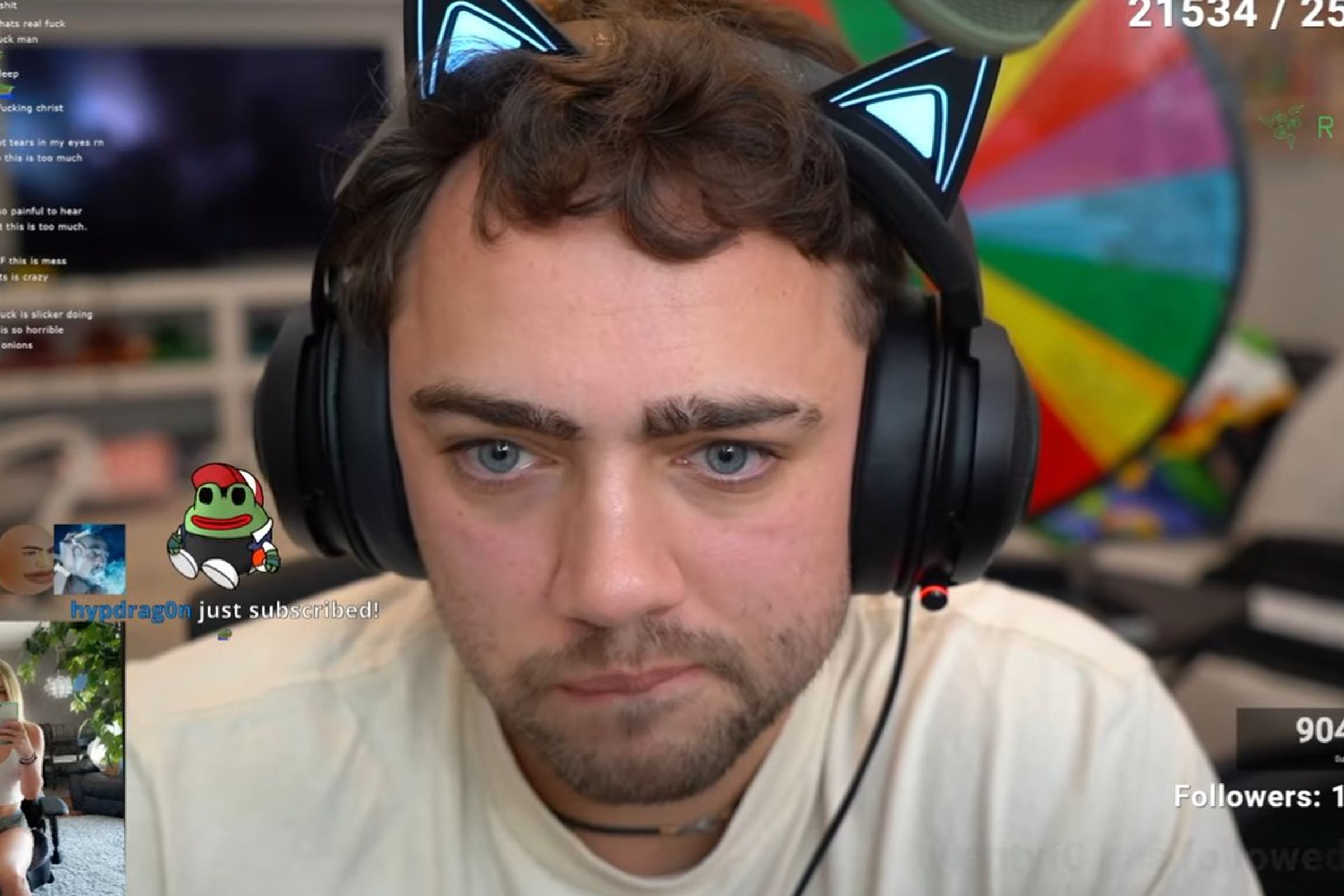 Image of Twitch streamer Mizkif, a brown-haired, fair-skinned male wearing a cream colored t-shirt and black headphones with neon blue cat ears poking up from the band.