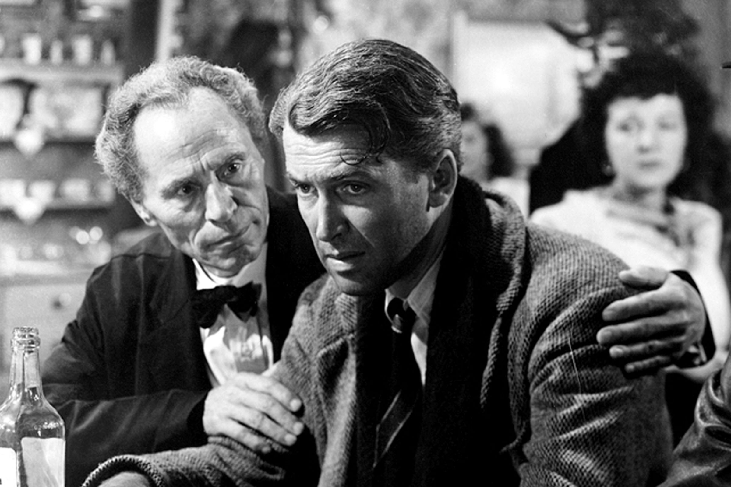 A man in a wool jacket and a scarf being comforted by another man in a wool jacket and a bowtie. Both men are standing at a brightly-lit bar or at a restaurant.