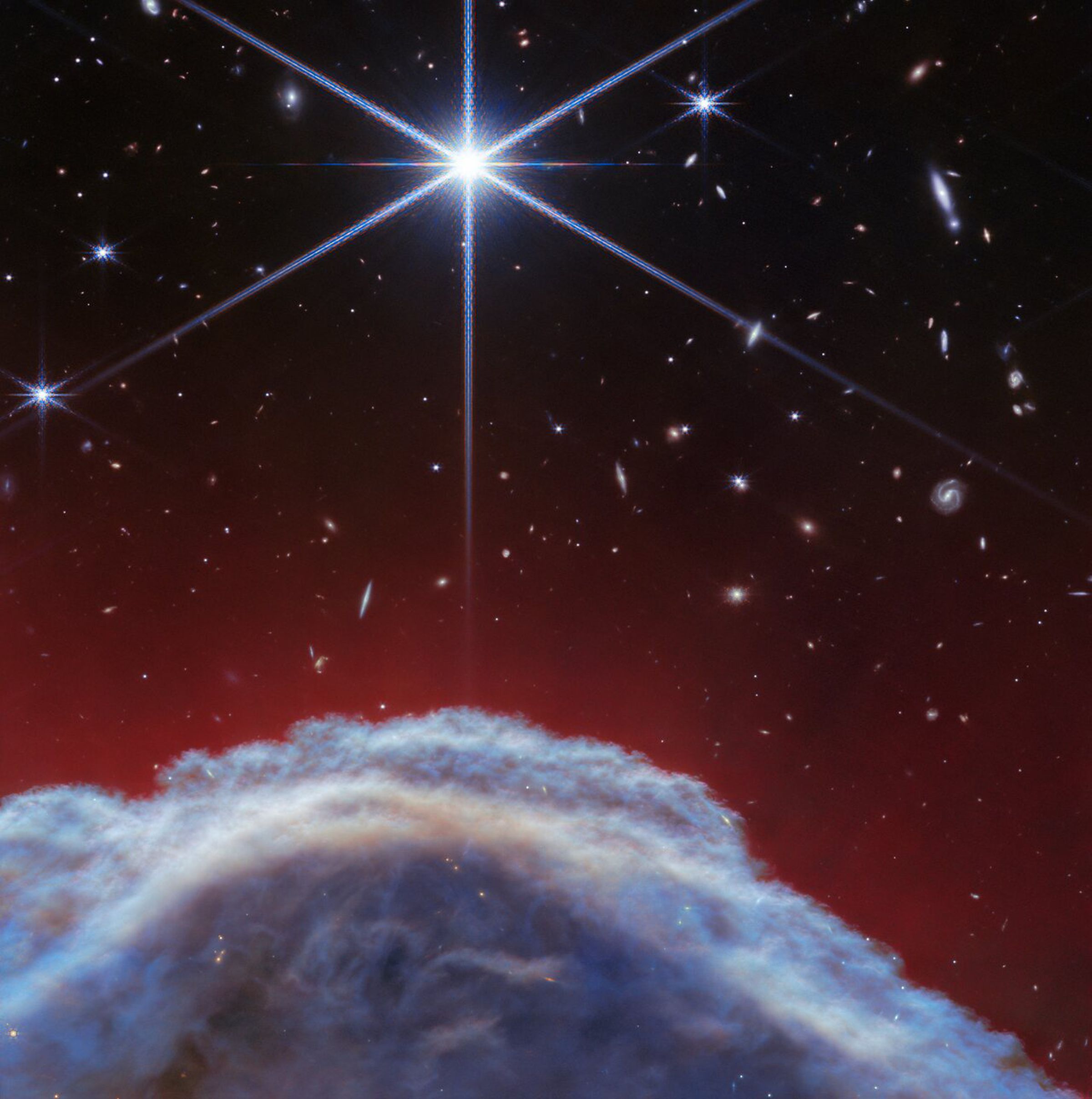 A color-shifted image of a portion of the Horsehead Nebula. The dust clouds are blue and brown, a reddish glow follows their contour, and stars and galaxies sit in the background.