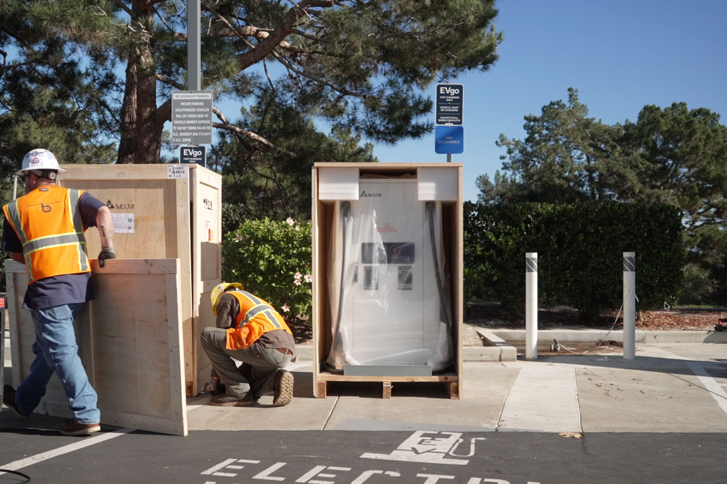 two workers in white hard hats and orange vests are opening large wooden crates in a parking lot in front of an EV charging spot. One crate reveals a charging station covered in plastic and foam protectors.