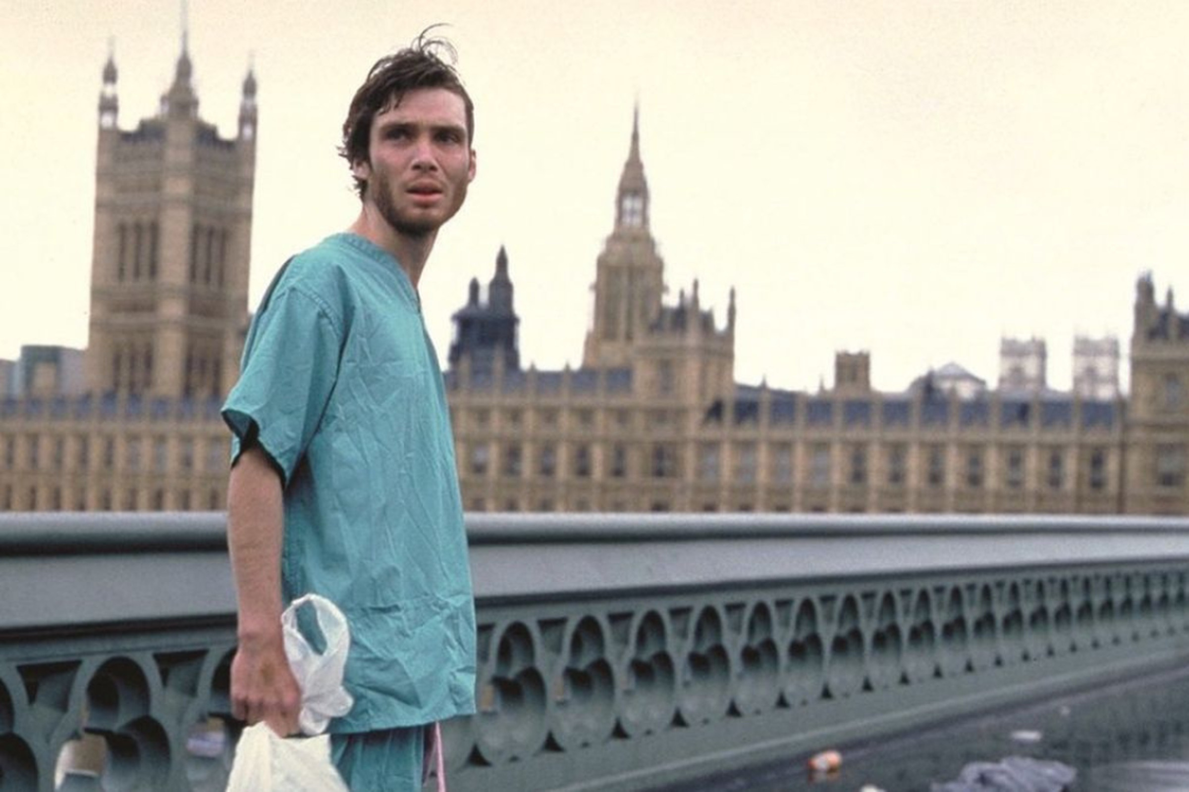 A man wearing hospital scrubs holding a plastic shopping back and looking out on a deserted London from the Westminster Bridge.