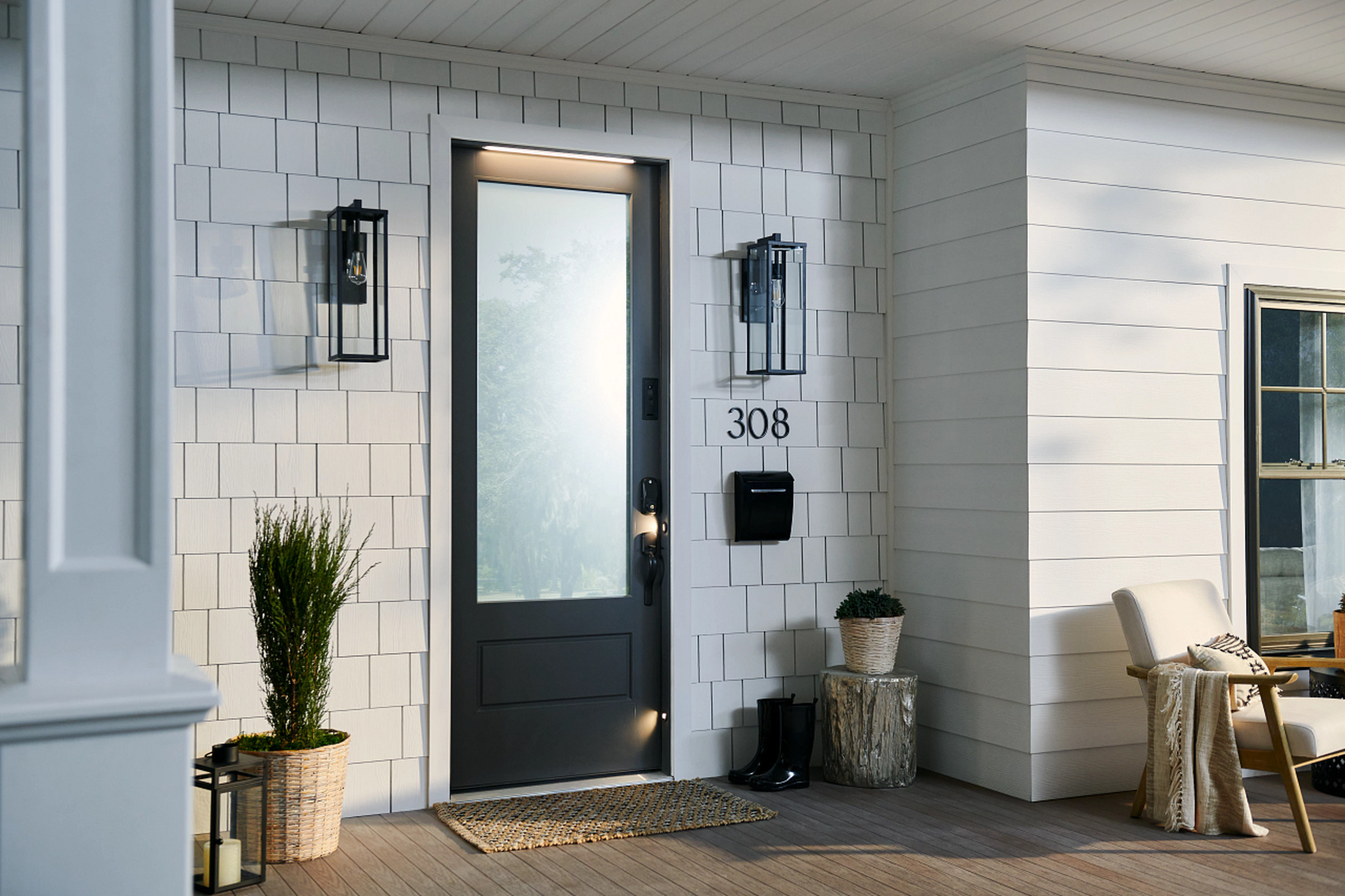 The front porch of a residential house with a Masonite M-Pwr smart door for its main entrance. The door is dark gray with a frosted glass panel, and it has a video doorbell, smart lock, and lighting built into it.