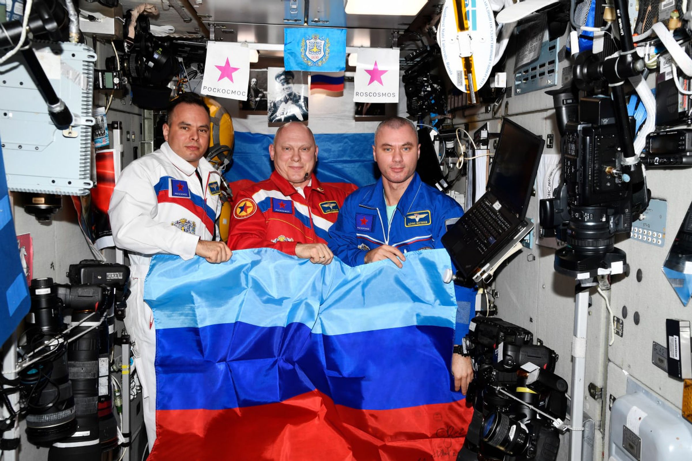 (L to R) Russian cosmonauts Sergey Korsakov, Oleg Artemyev, and Denis Matveev pose with the flag of the Luhansk People’s Republic