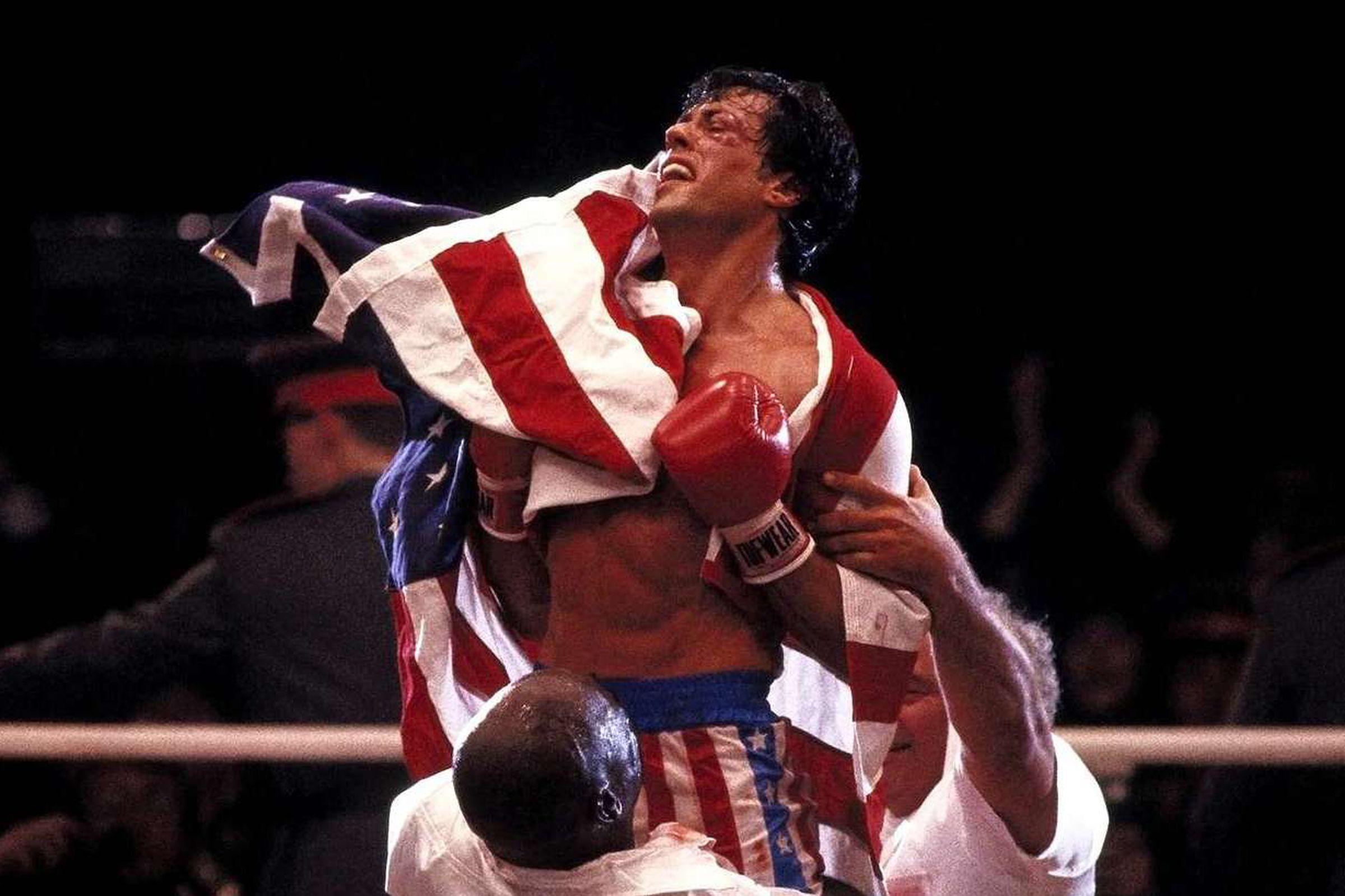 A bruised boxer wrapped in an American flag being housed up by two other men wearing white shirts.