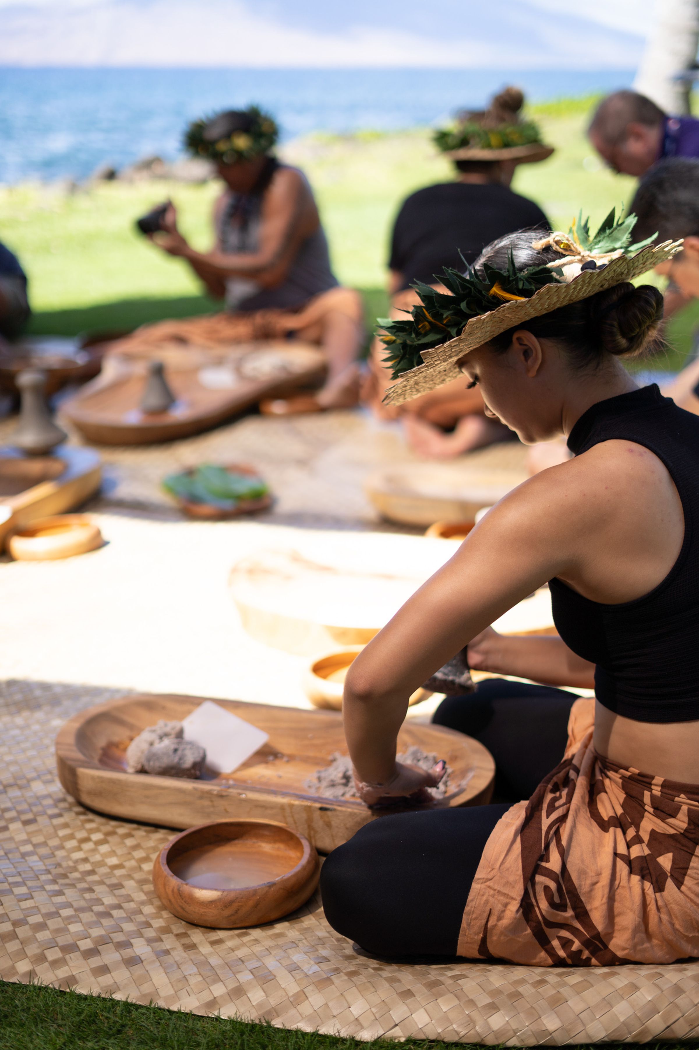 Woman in traditional Hawaiian dress seated on a mat pounding starch into poi.