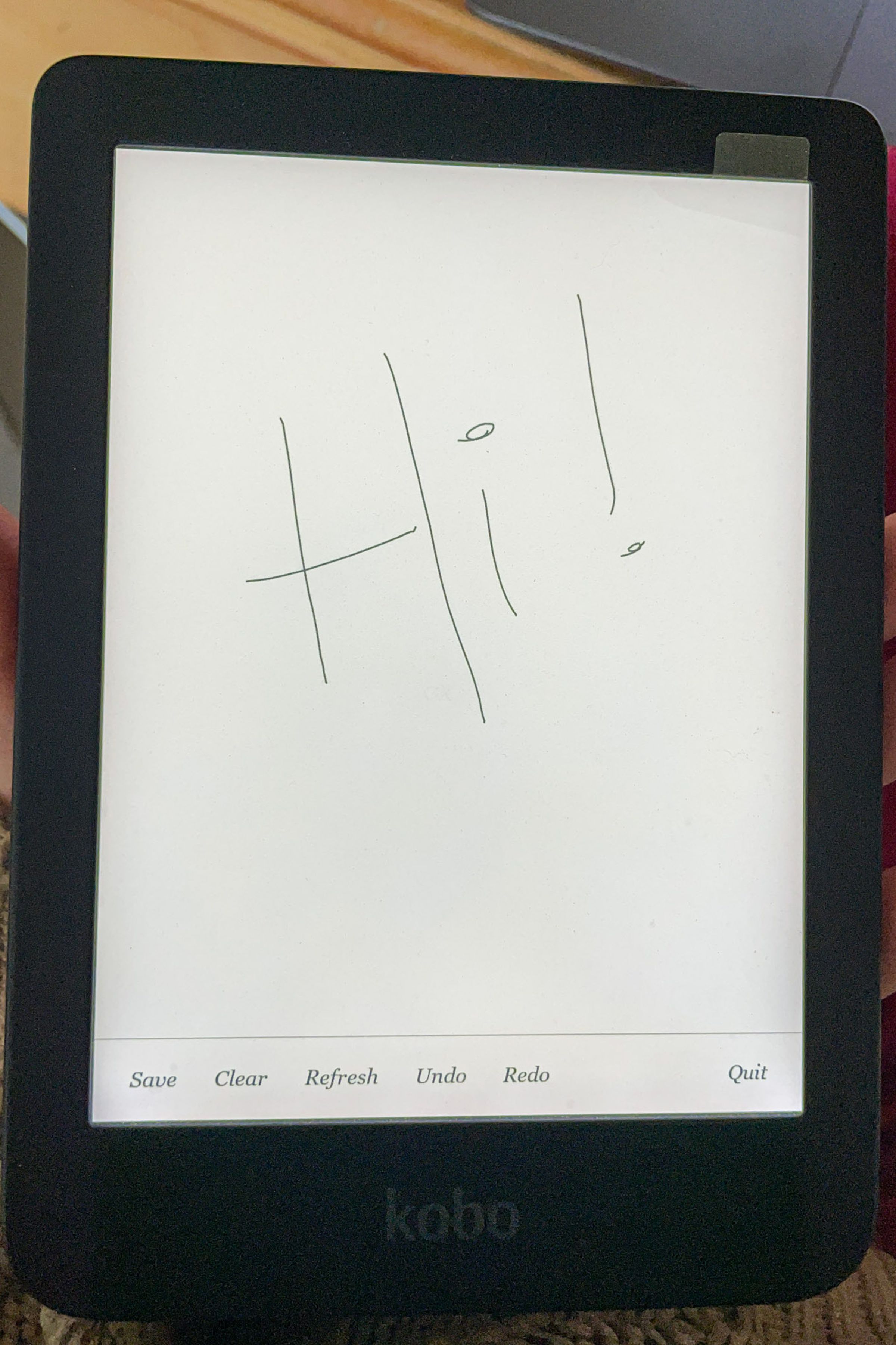 A hand holding up the Kobo Clara 2E. Its screen is on and open to the experimental sketchbook app, where the word “Hi” is written in handwriting.