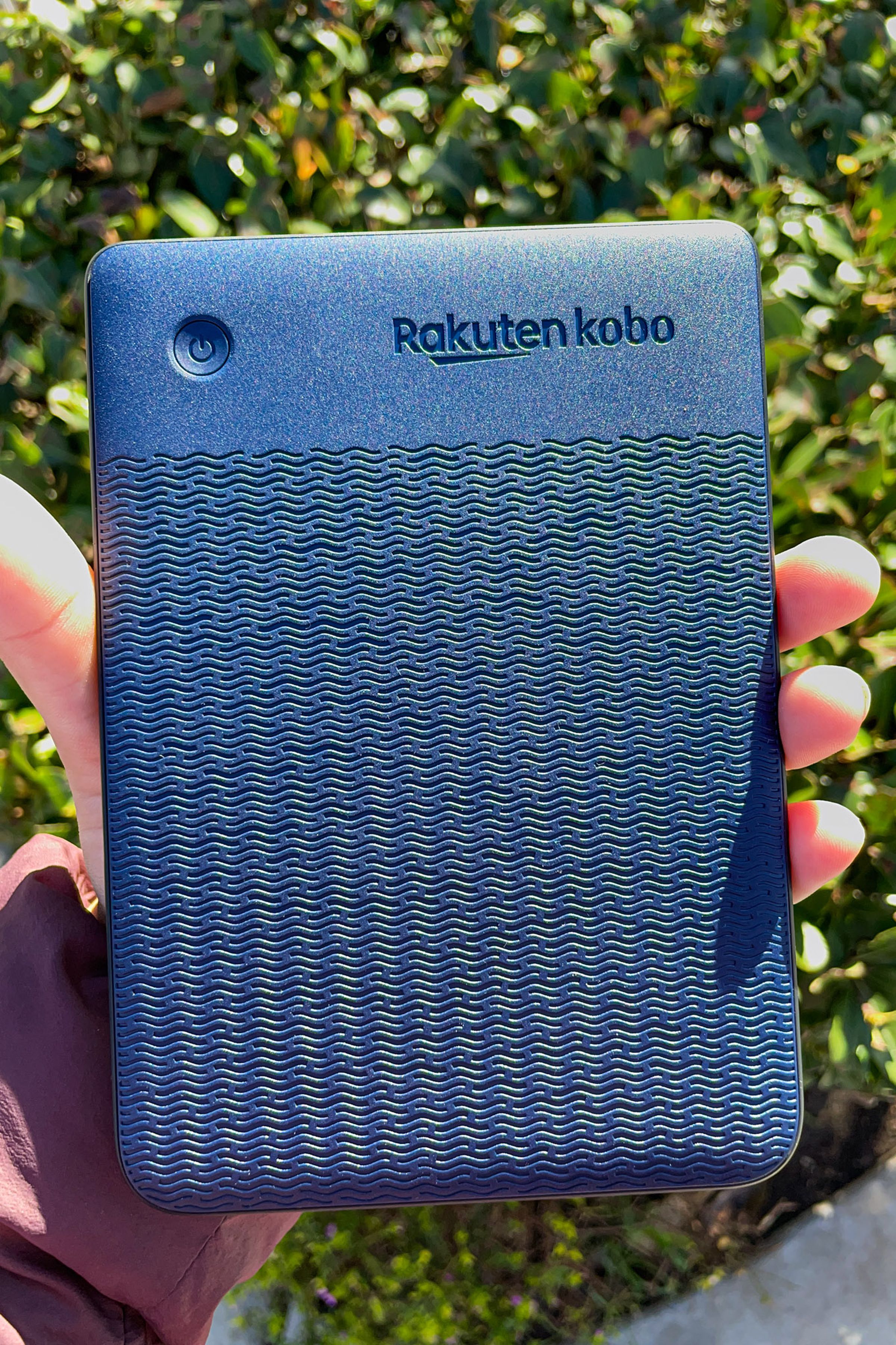 A hand holding up the Kobo Clara 2E turned around so its denim blue back, wavy pattern, and power button is on display.