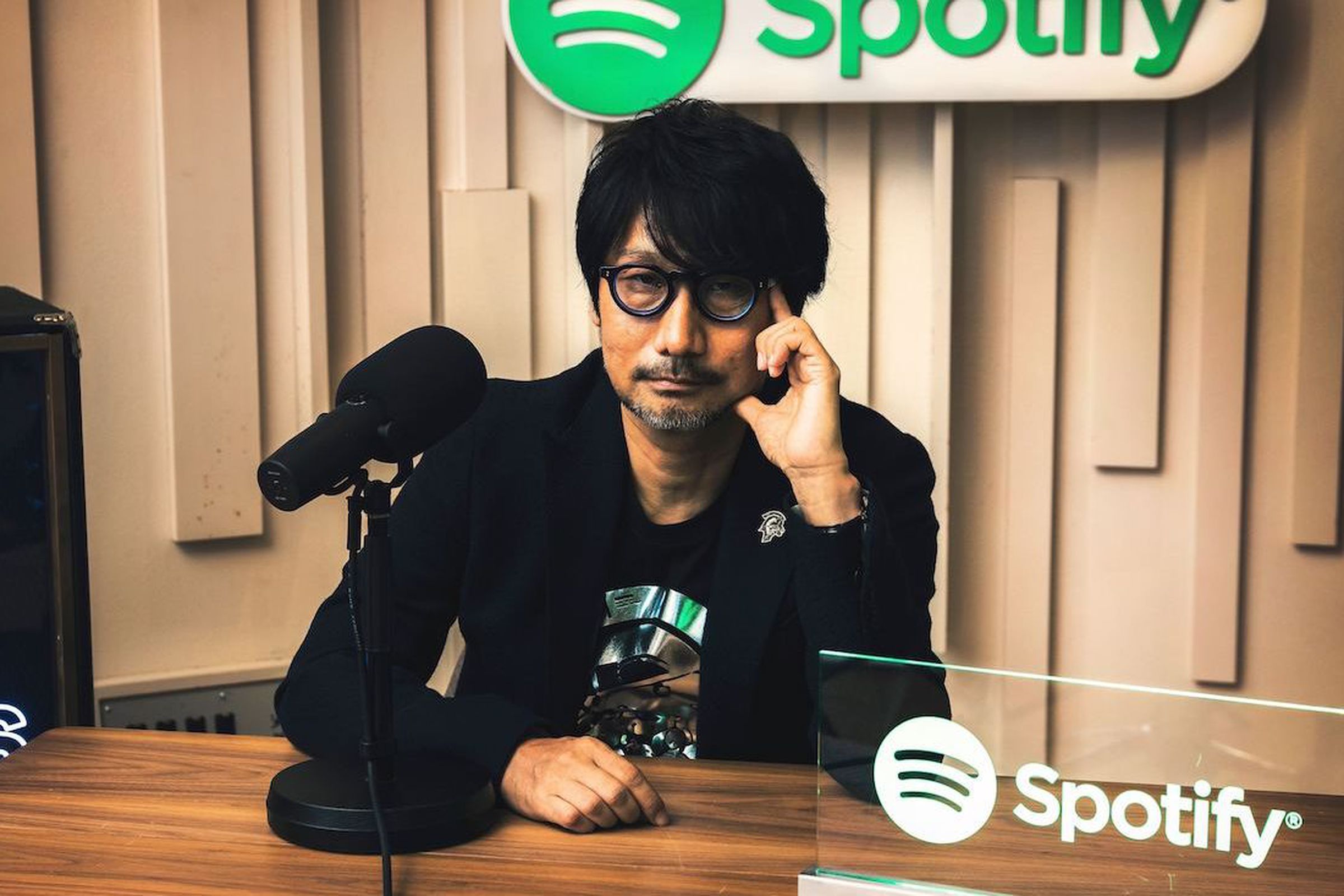 Hideo Kojima leaning thoughtfully at a desk with a microphone next to him with the Spotify logo hanging in the background