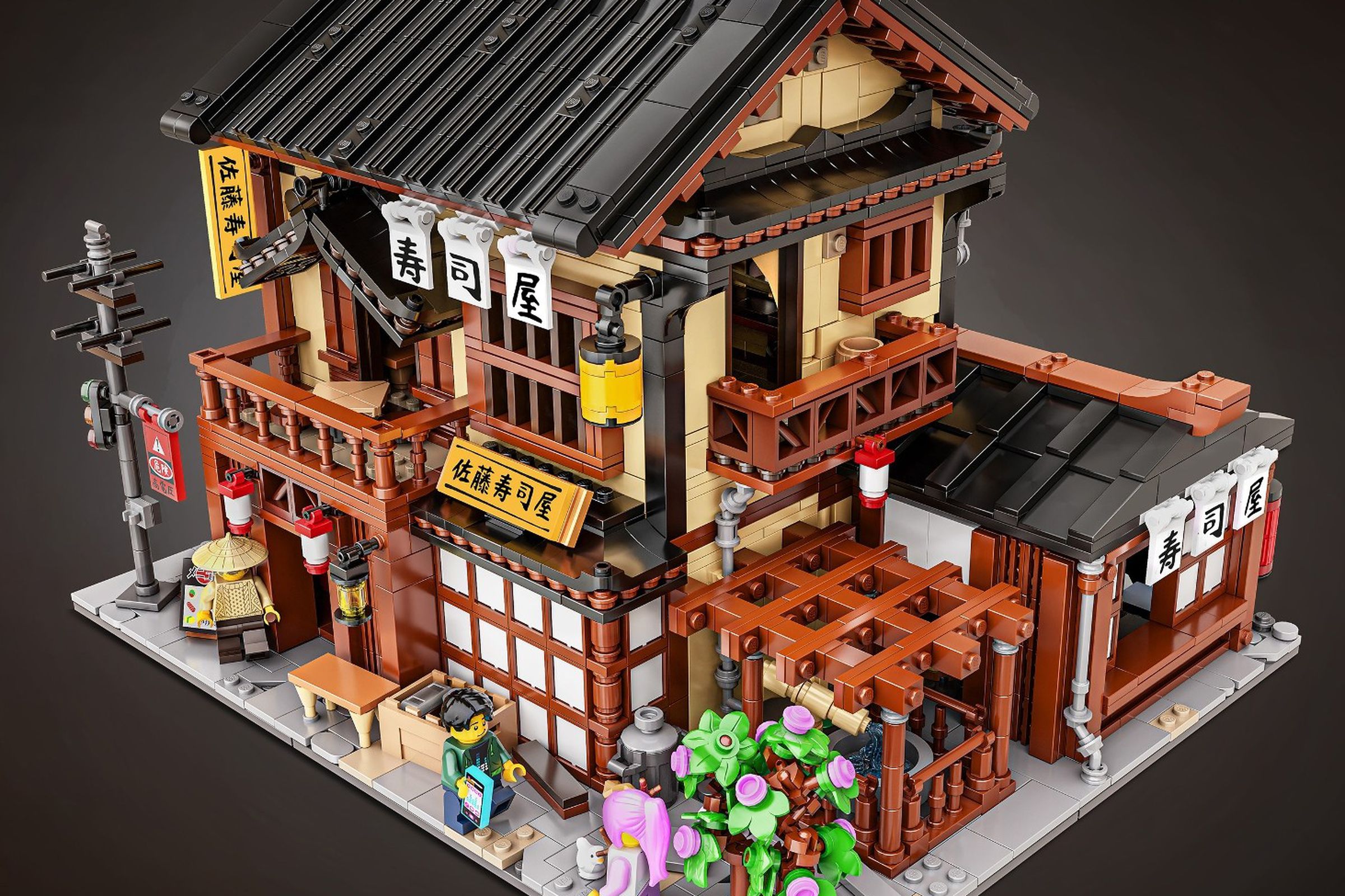 A traditional looking Japanese building made entirely out of Lego, with a fountain, flowers, two stories, entranceways and signs to the Sushi restaurant.