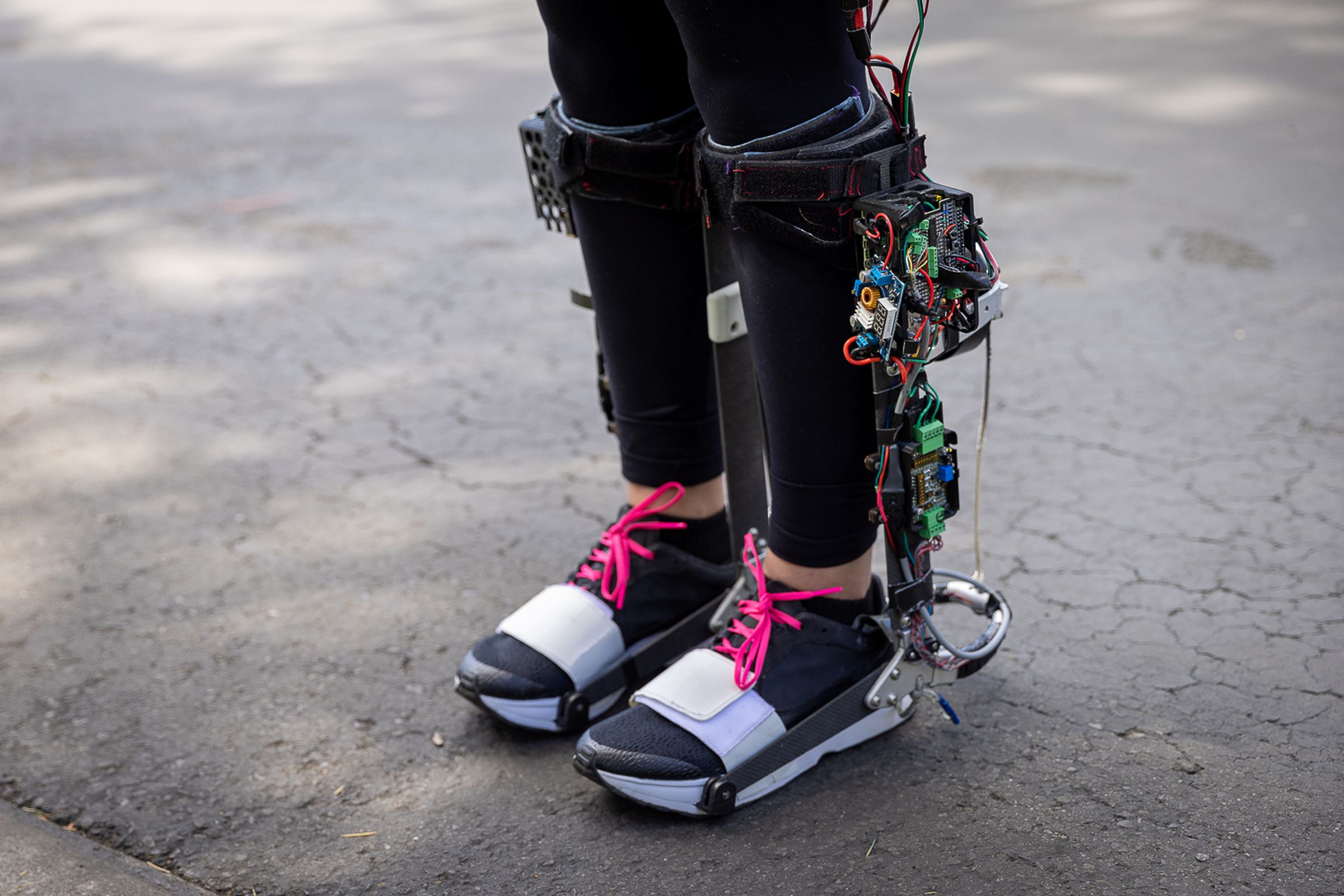 An image of someone’s legs from the knee down with a robotic exoskeleton attached at the feet and below the knee. The exoskeleton is exposed, colorful wires and circuit boards.