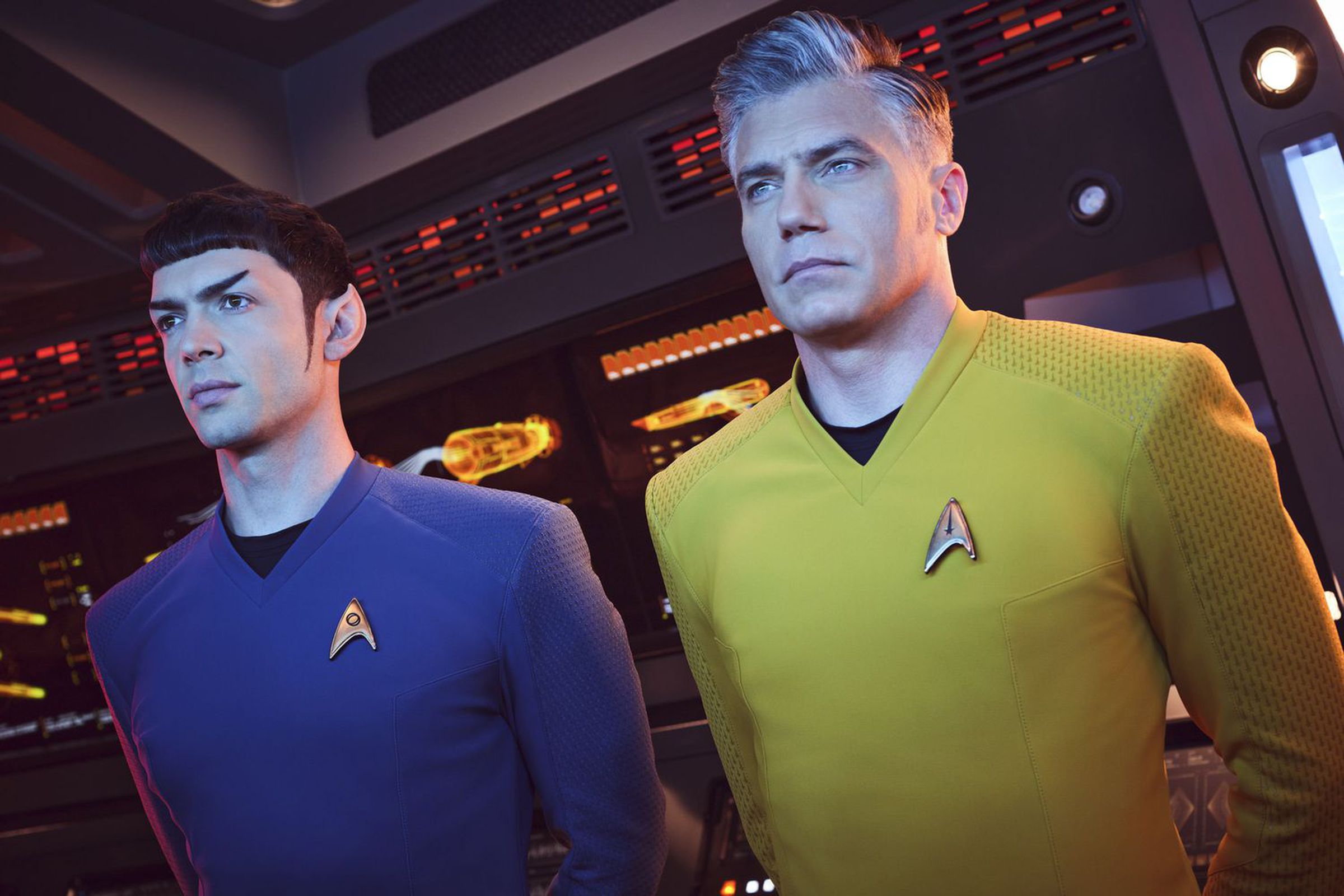 Two men, one wearing a blue shirt, and the other wearing a yellow short, standing with their arms behind their backs in a futuristic control room.