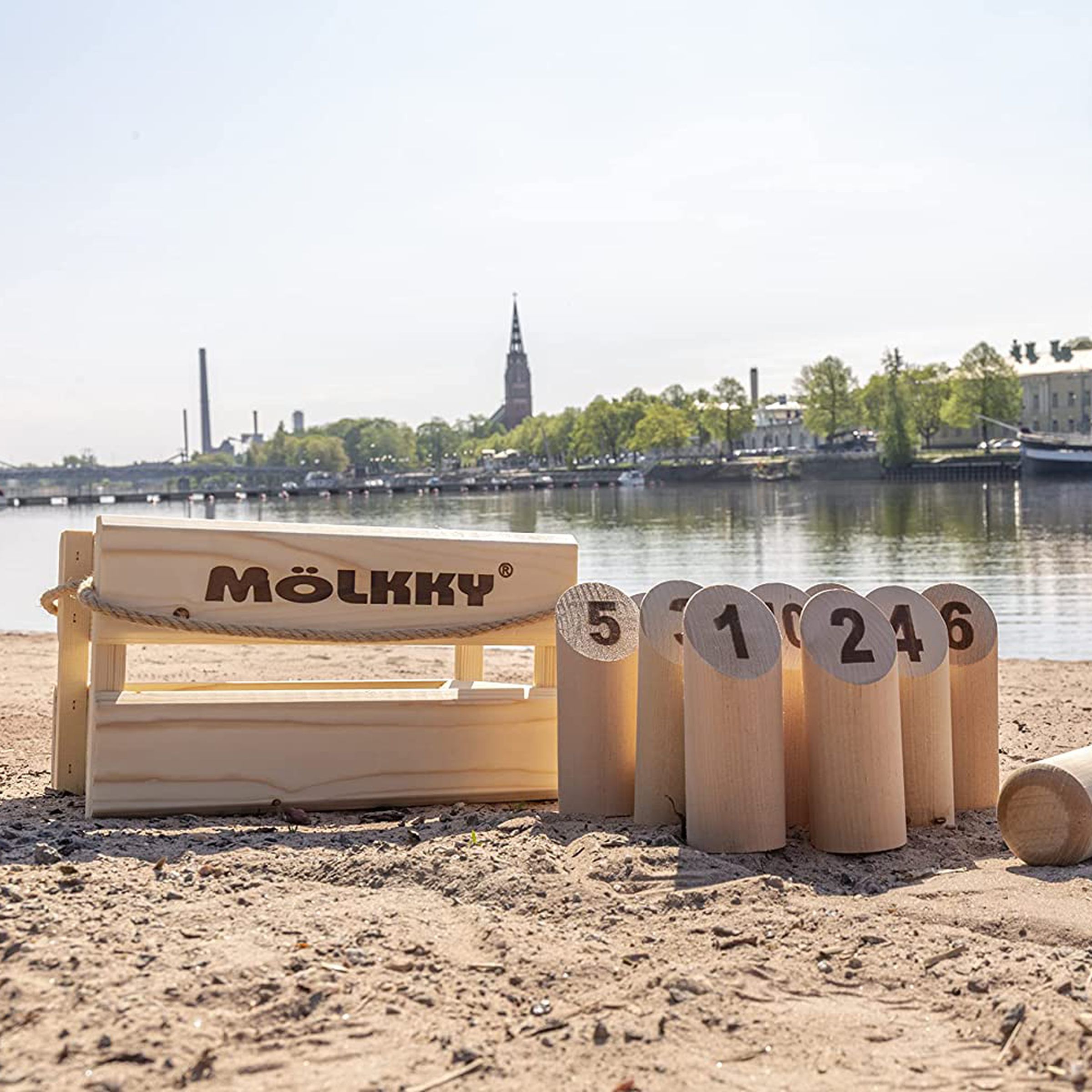 Molkky game with a wooden, several numbered pins, and a wooden dowel, on a beach with a town in the background.