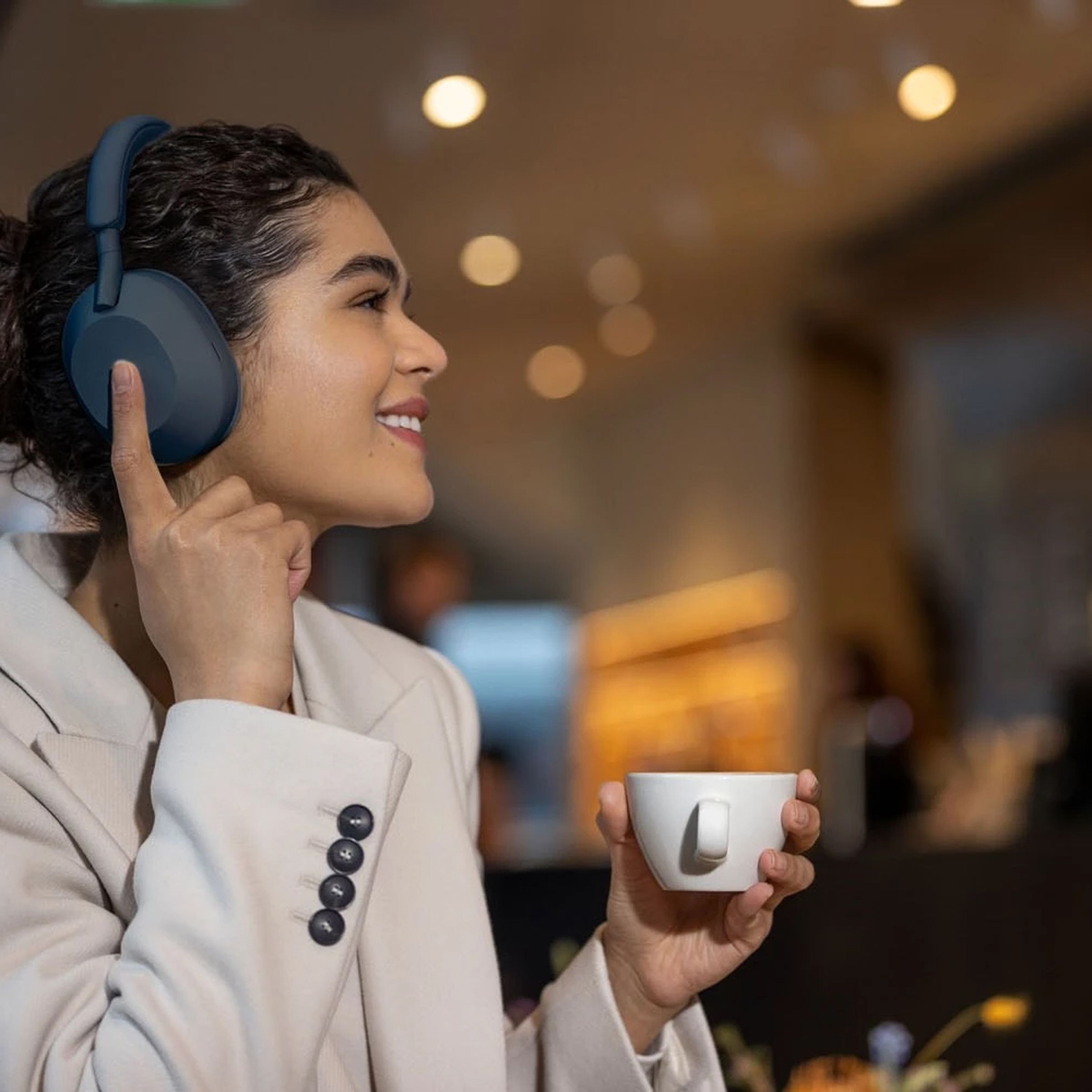 A woman in a suit wearing a pair of Sony’s WH-1000XM5 headphones while drinking coffee and adjusting playback.
