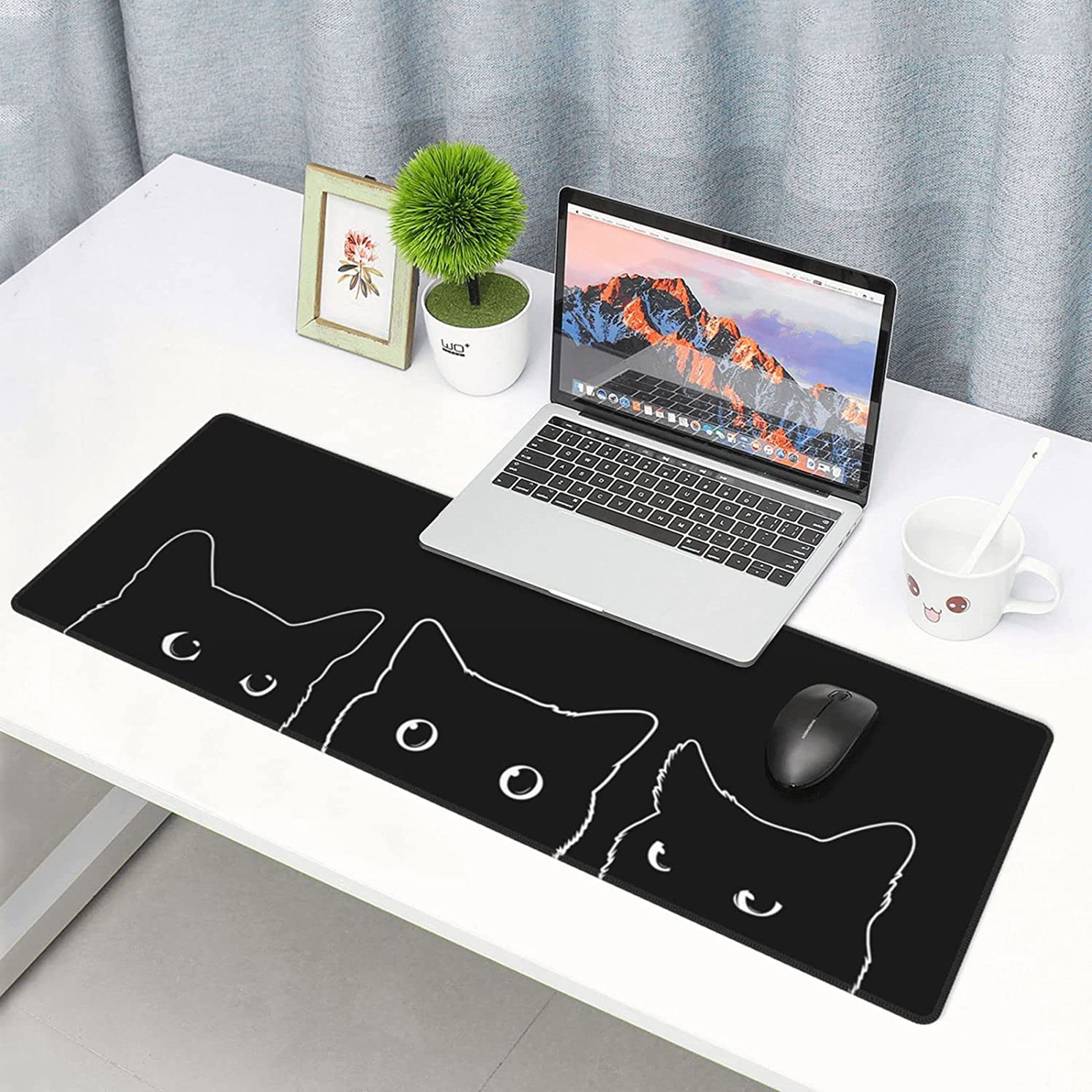 Um longo mouse pad preto com os contornos de três gatos em uma mesa branca com um laptop, uma planta e uma pequena foto emoldurada.