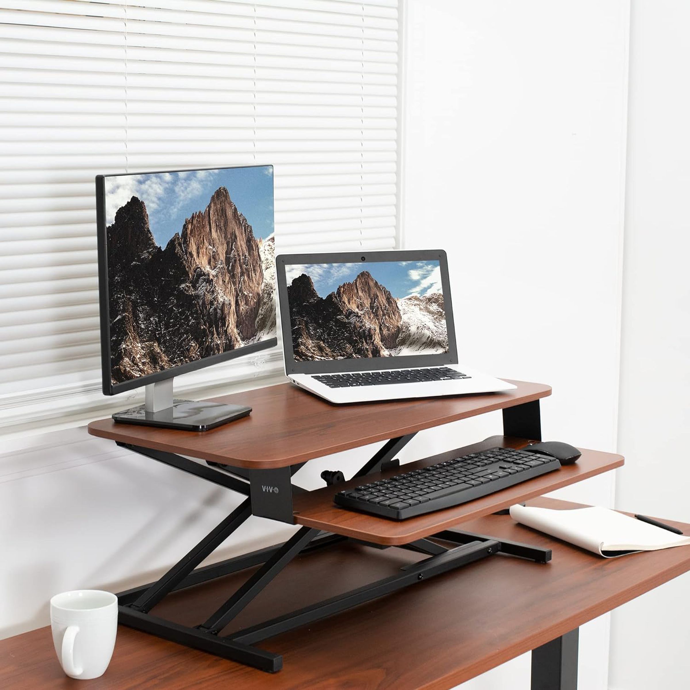 A two-level stand on top of a desk holding a monitor, a laptop, and a keyboard.