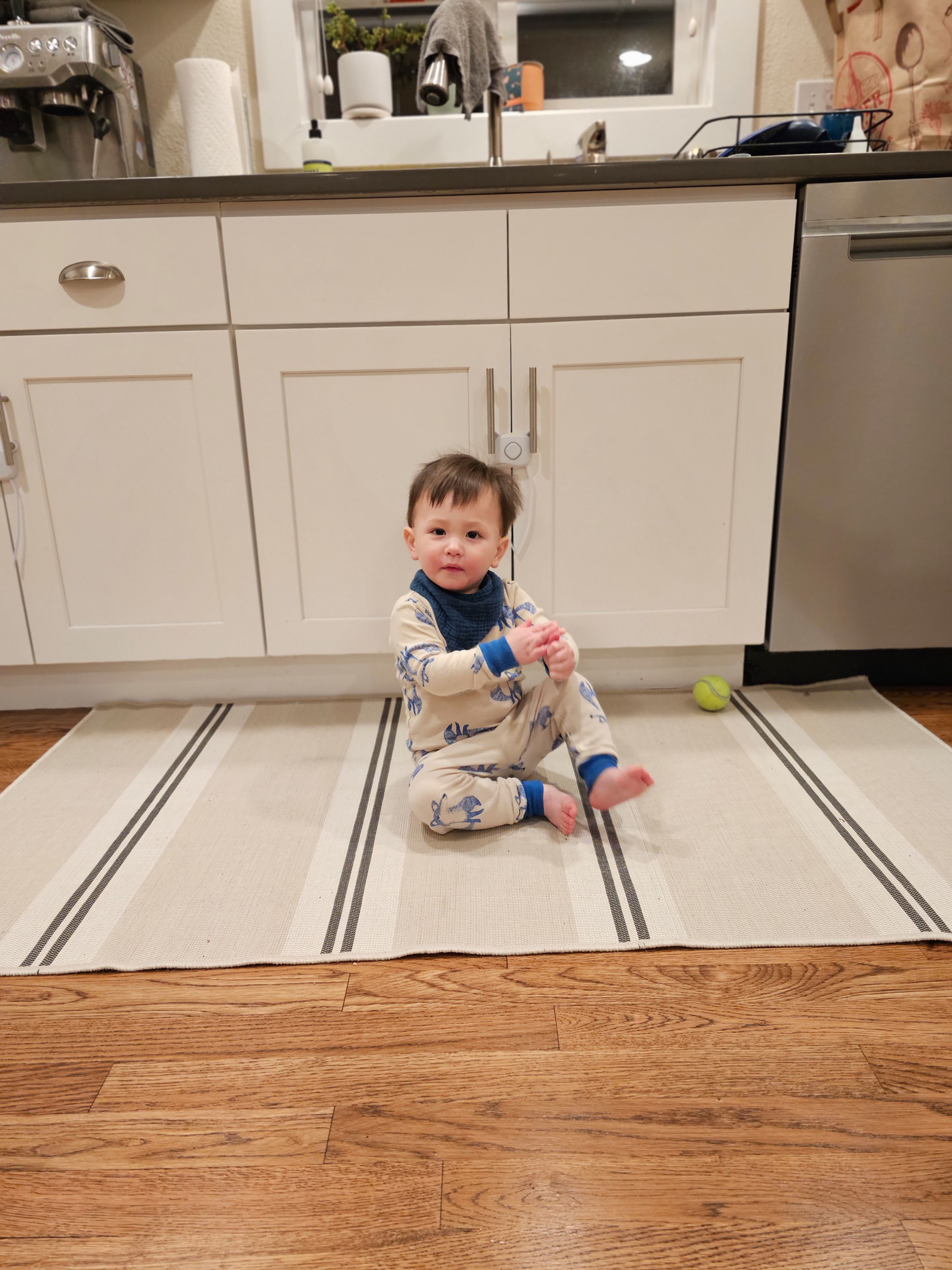 Picture of a toddler sitting on a kitchen rug looking at the camera.