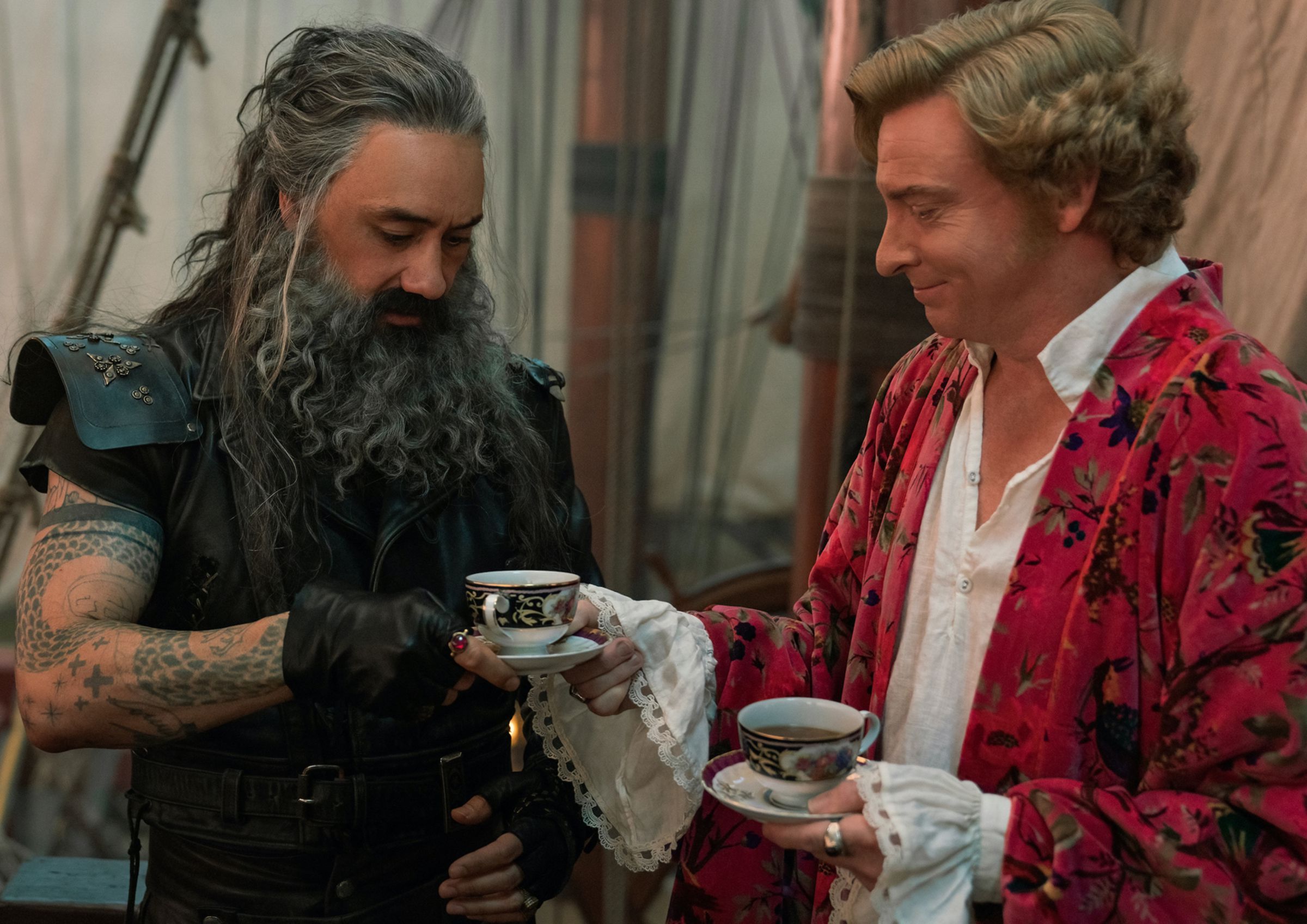 A roughhewn pirate with a massive beard taking a cup of tea from a fey man in a dressing gown and a night coat.