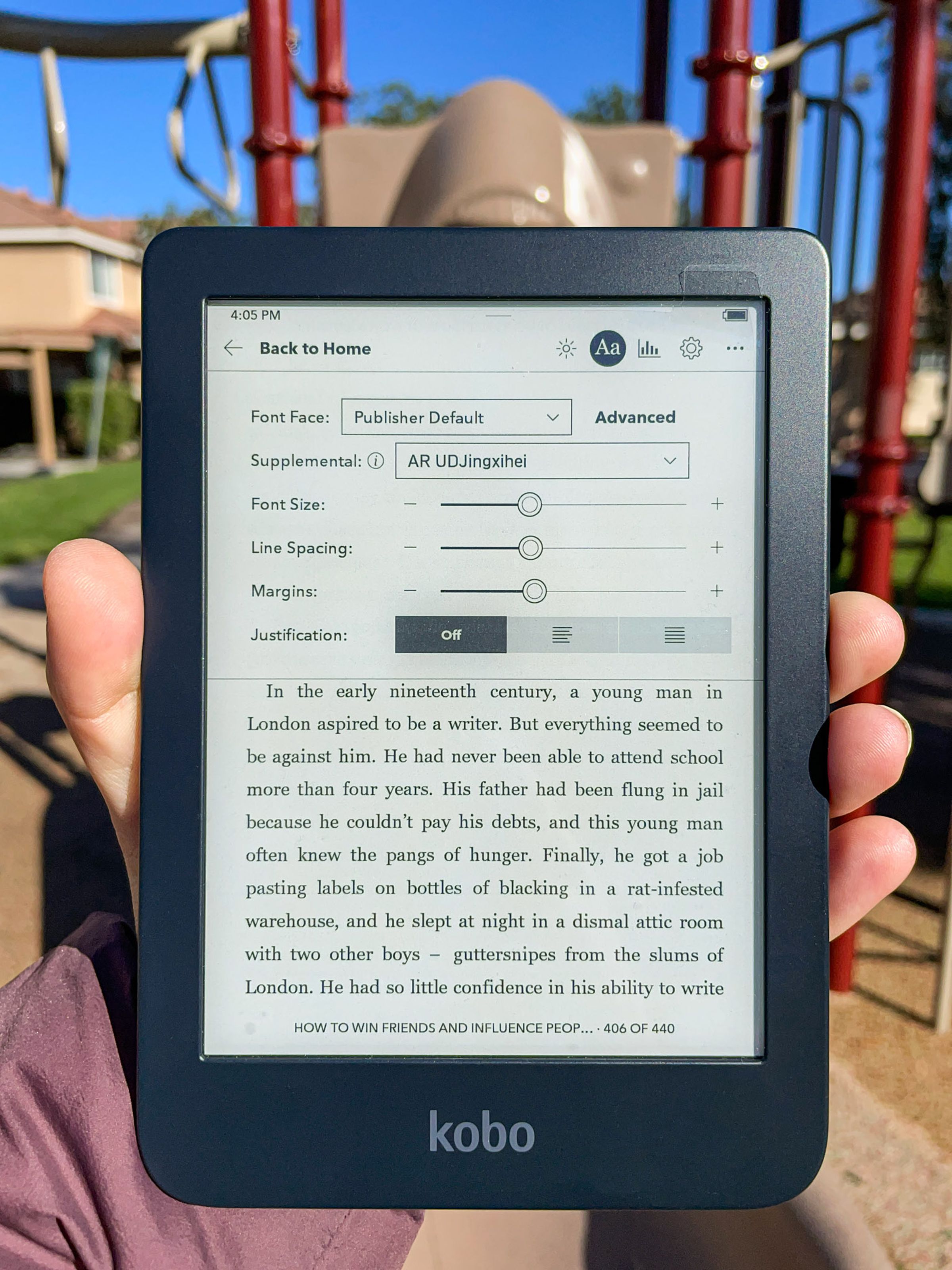 A hand holding up the Kobo Clara 2E. Its screen is on and open to a page in an ebook, while a menu is on display with options to adjust the font size, line spacing, and more using sliders.