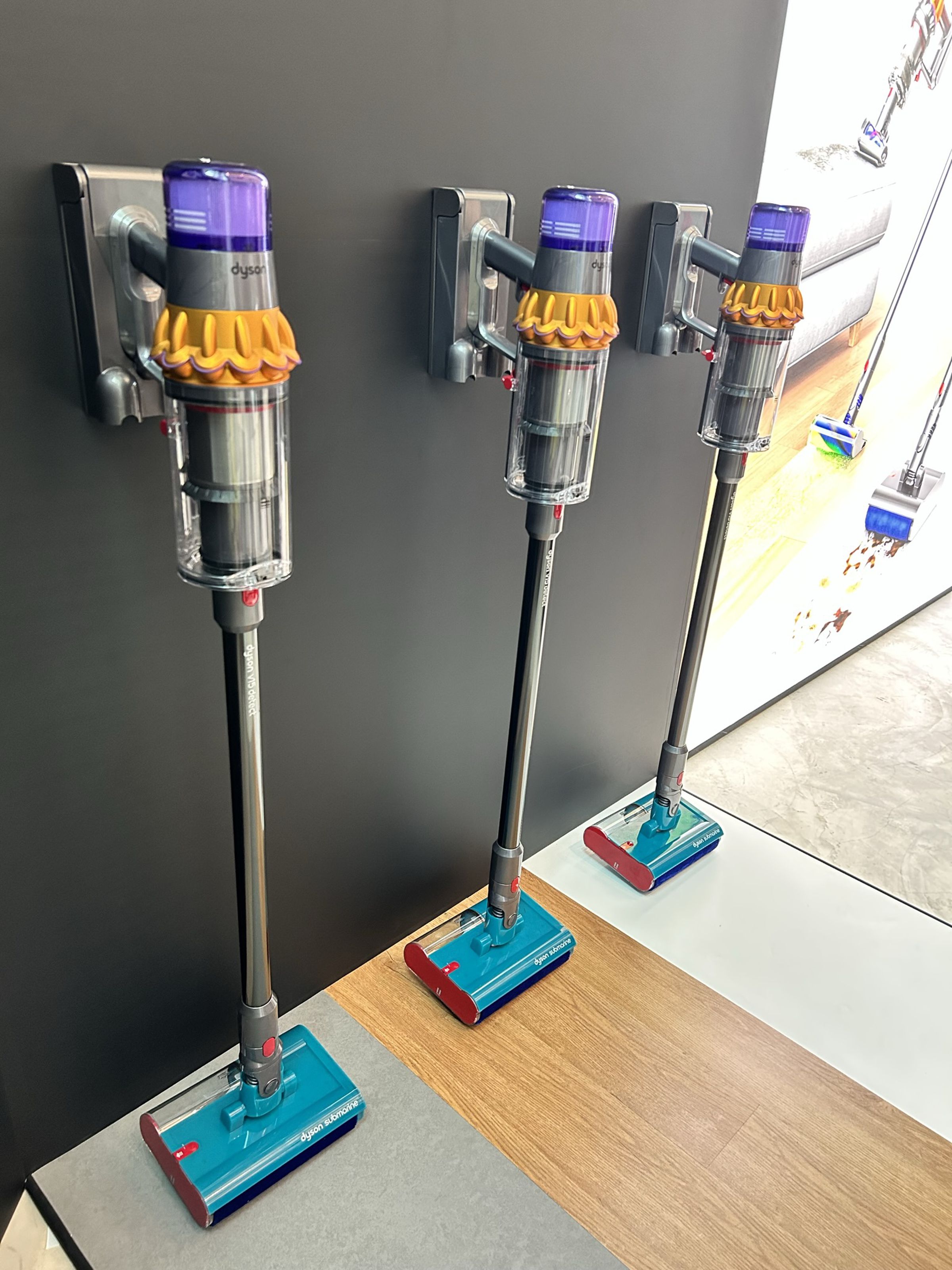 Three identical stick vacs mounted to a wall. They are mostly a drab gray, but the new Submarine heads are brightly colored turquoise and red.