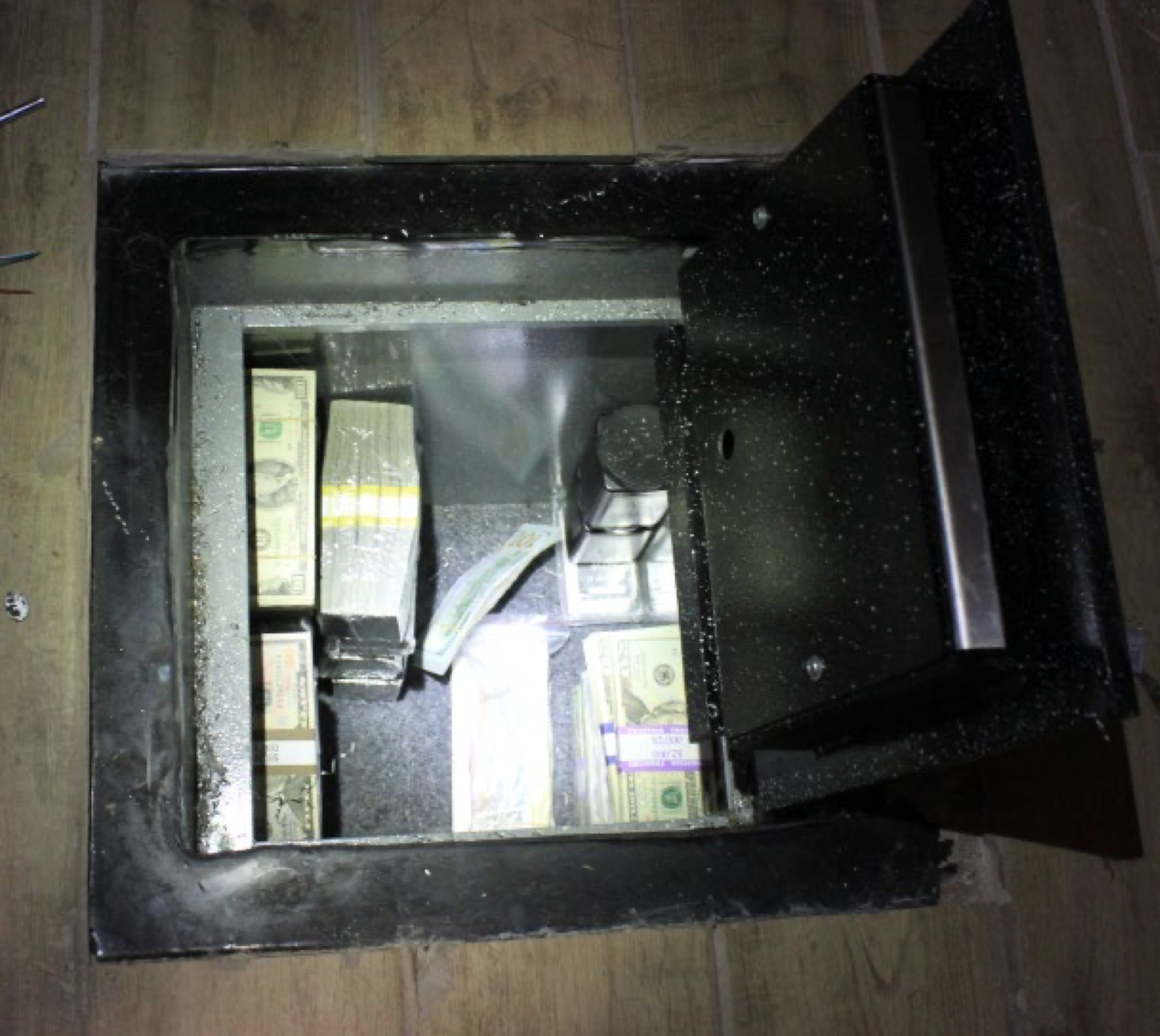 Image of a safe embedded in a wooden floor, containing cash and two metallic objects.