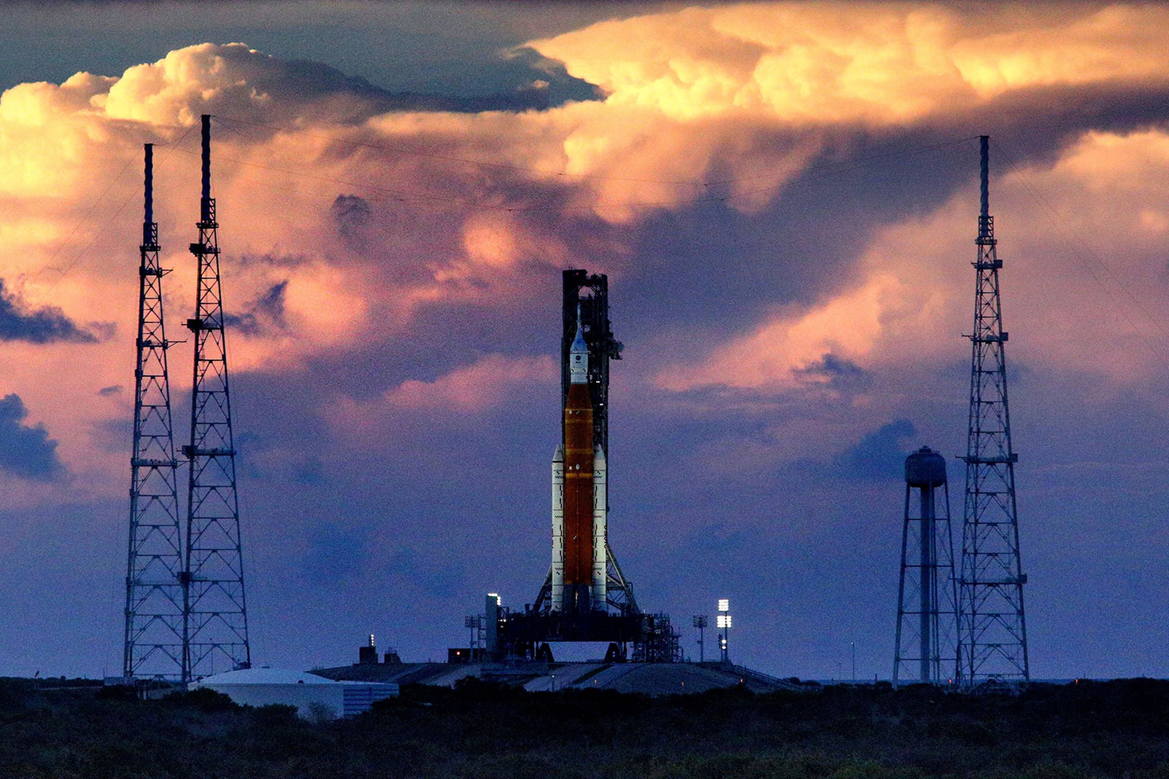 A rocket sits on the launchpad at the center of the image with sunset-lit clouds looming in the background