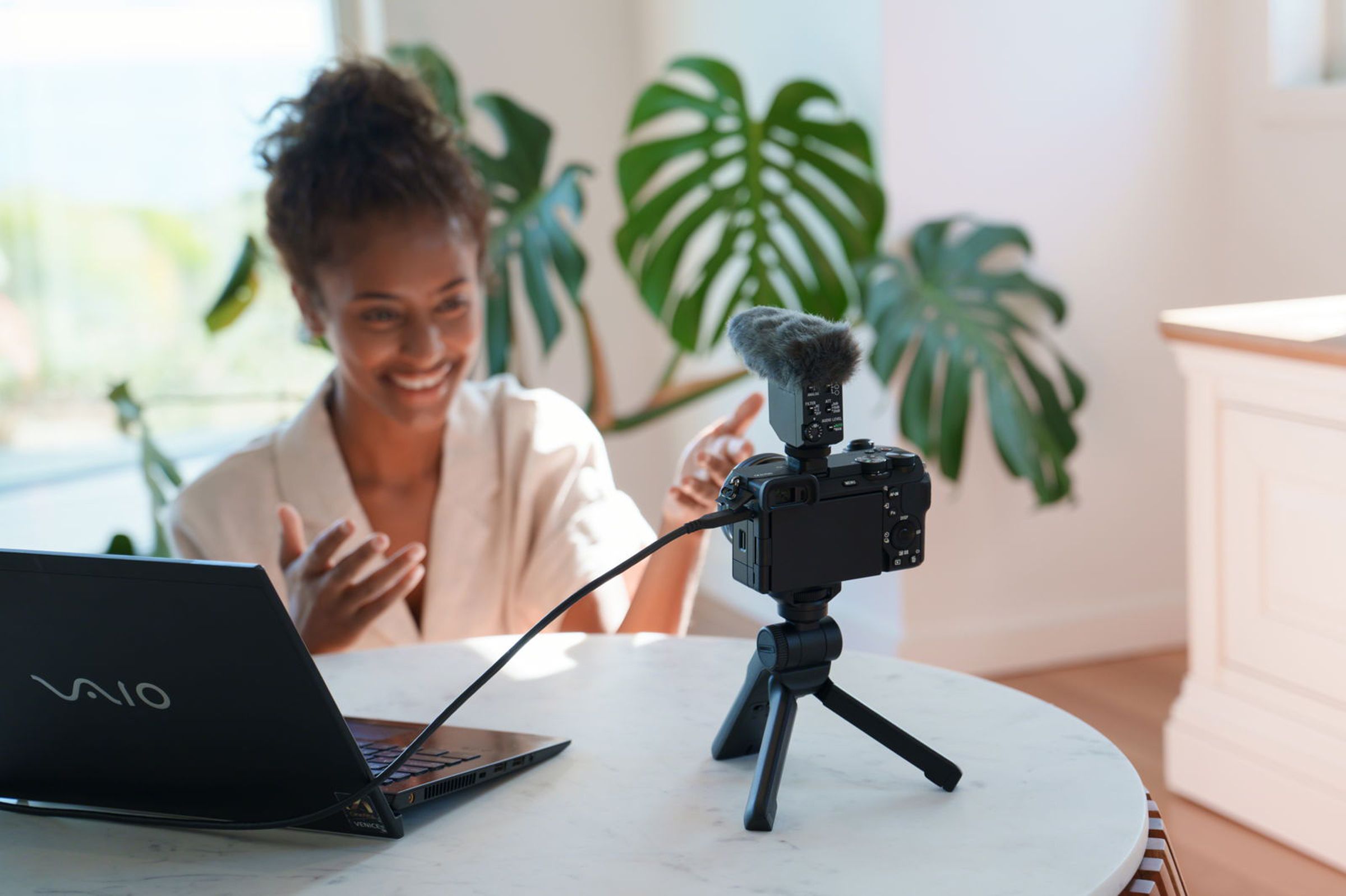 Shot showing a person sitting at a table, smiling into the Sony A6700, which sits on a small tabletop tripod stand and has a microphone attached. A laptop sits on the table to the left, and the camera is attached to it with a cable.