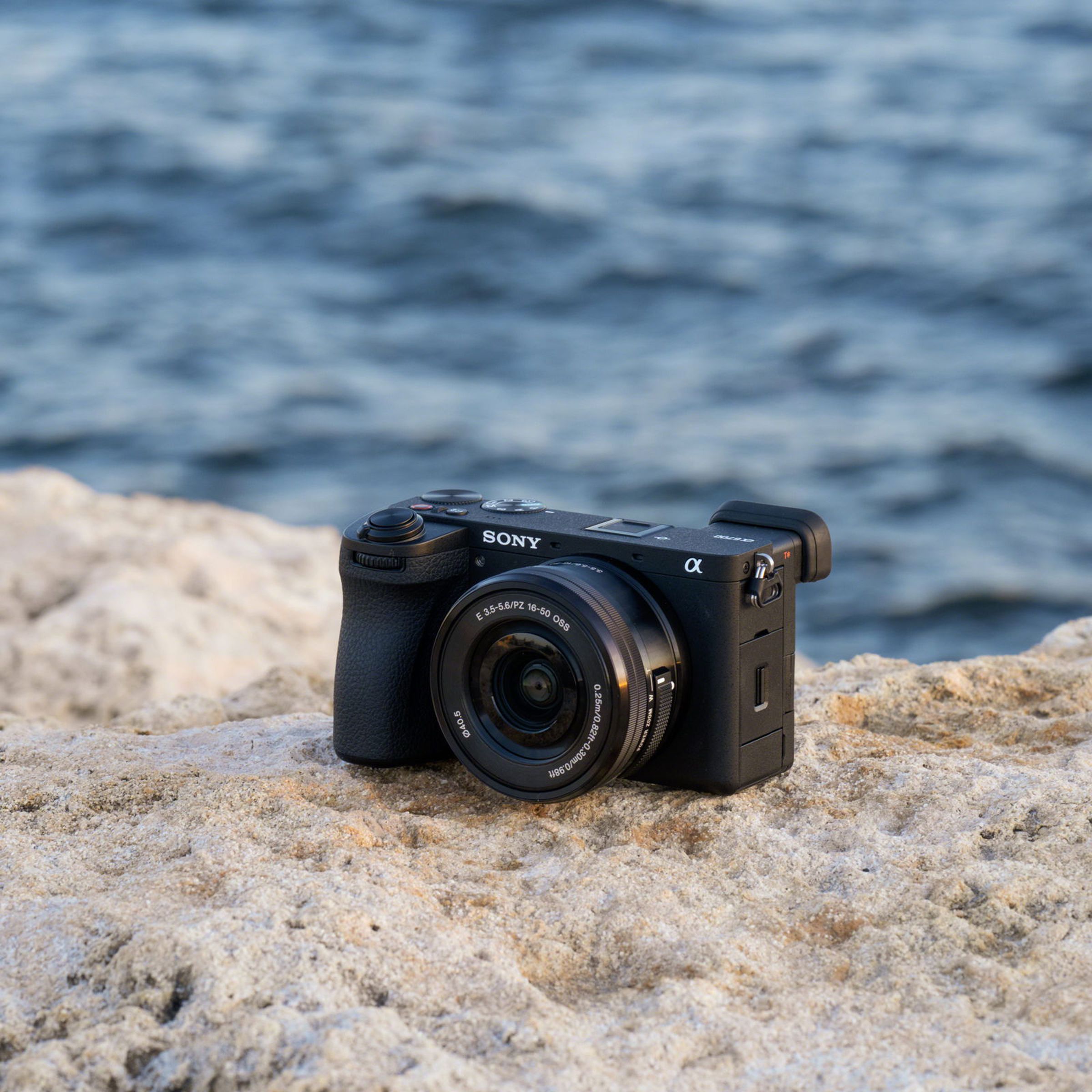 A three-quarter shot of the Sony A6700 sitting on a porous, beige rock near what appears to be ocean water.