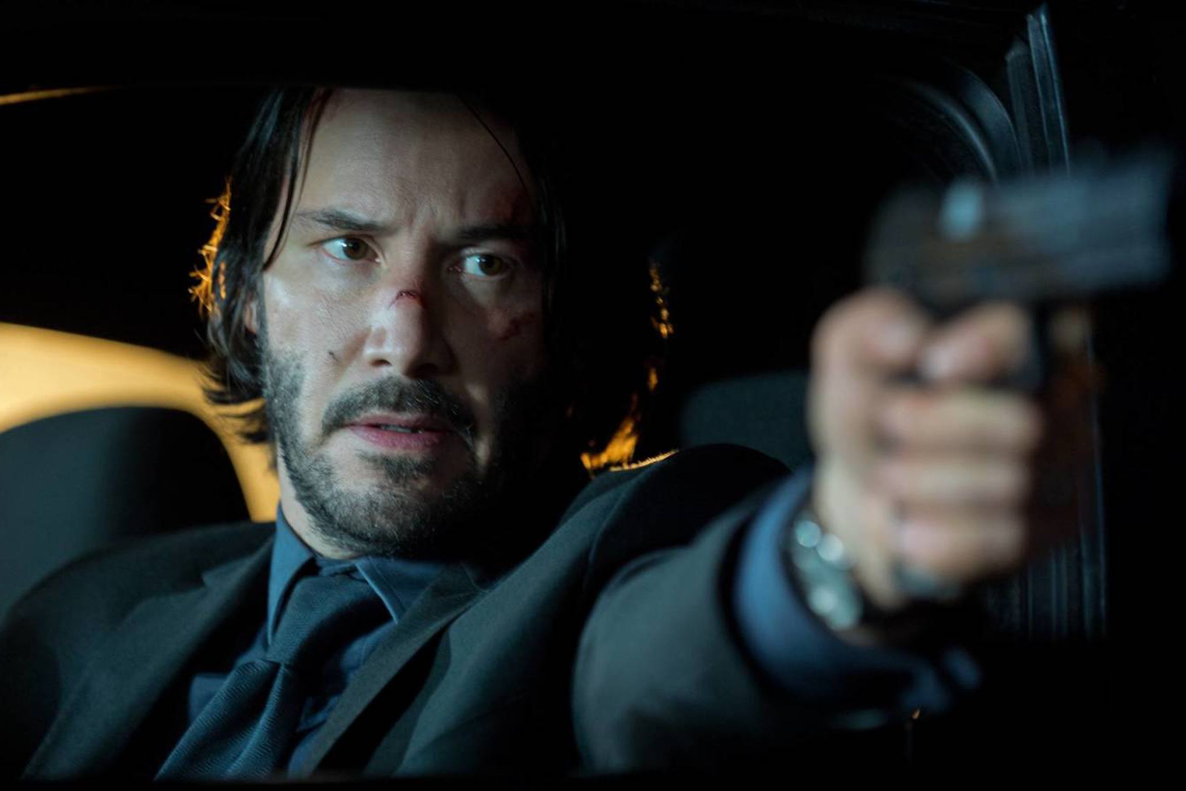 A man in a suit sitting in a car and pointing a gun out the window.