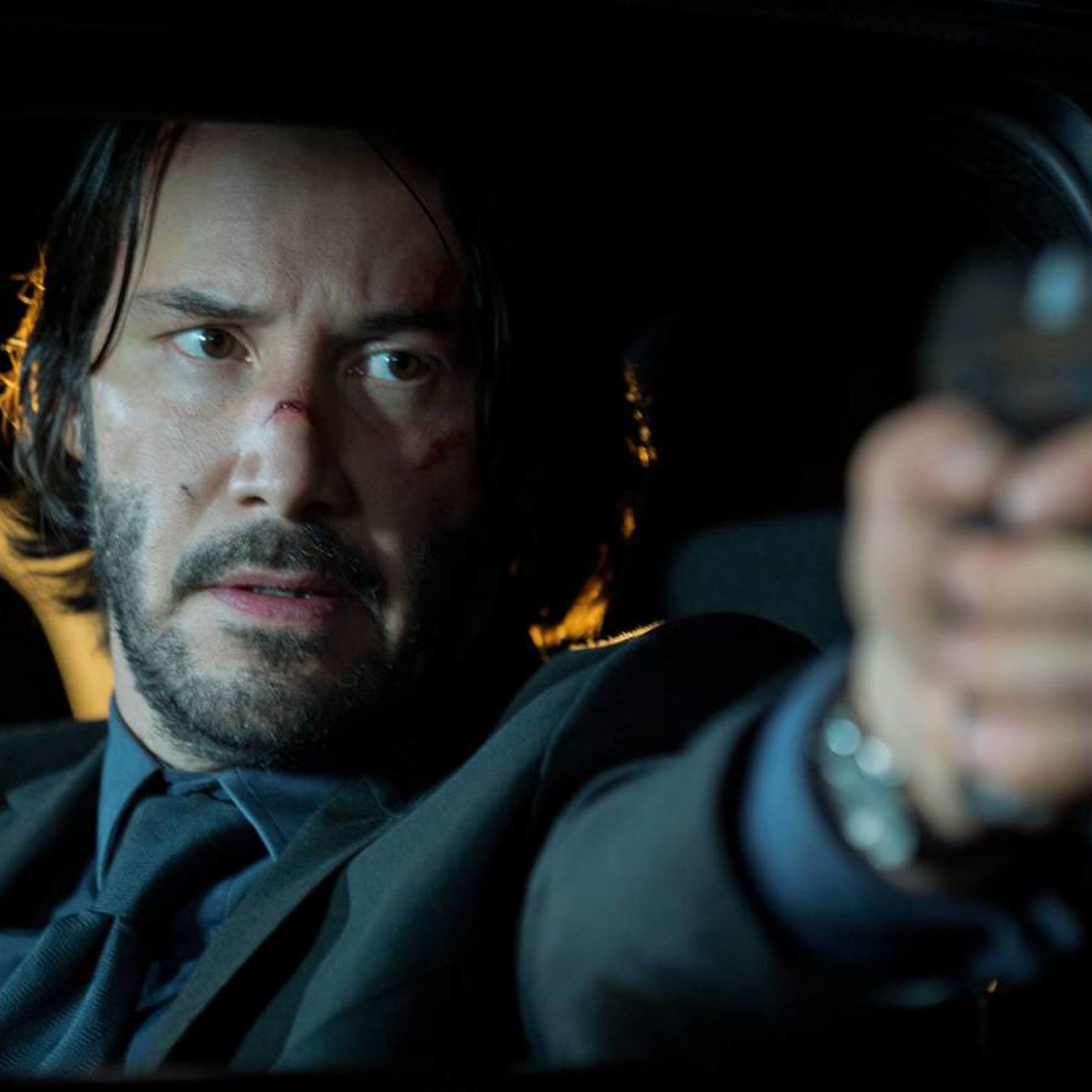 A man in a suit sitting in a car and pointing a gun out the window.