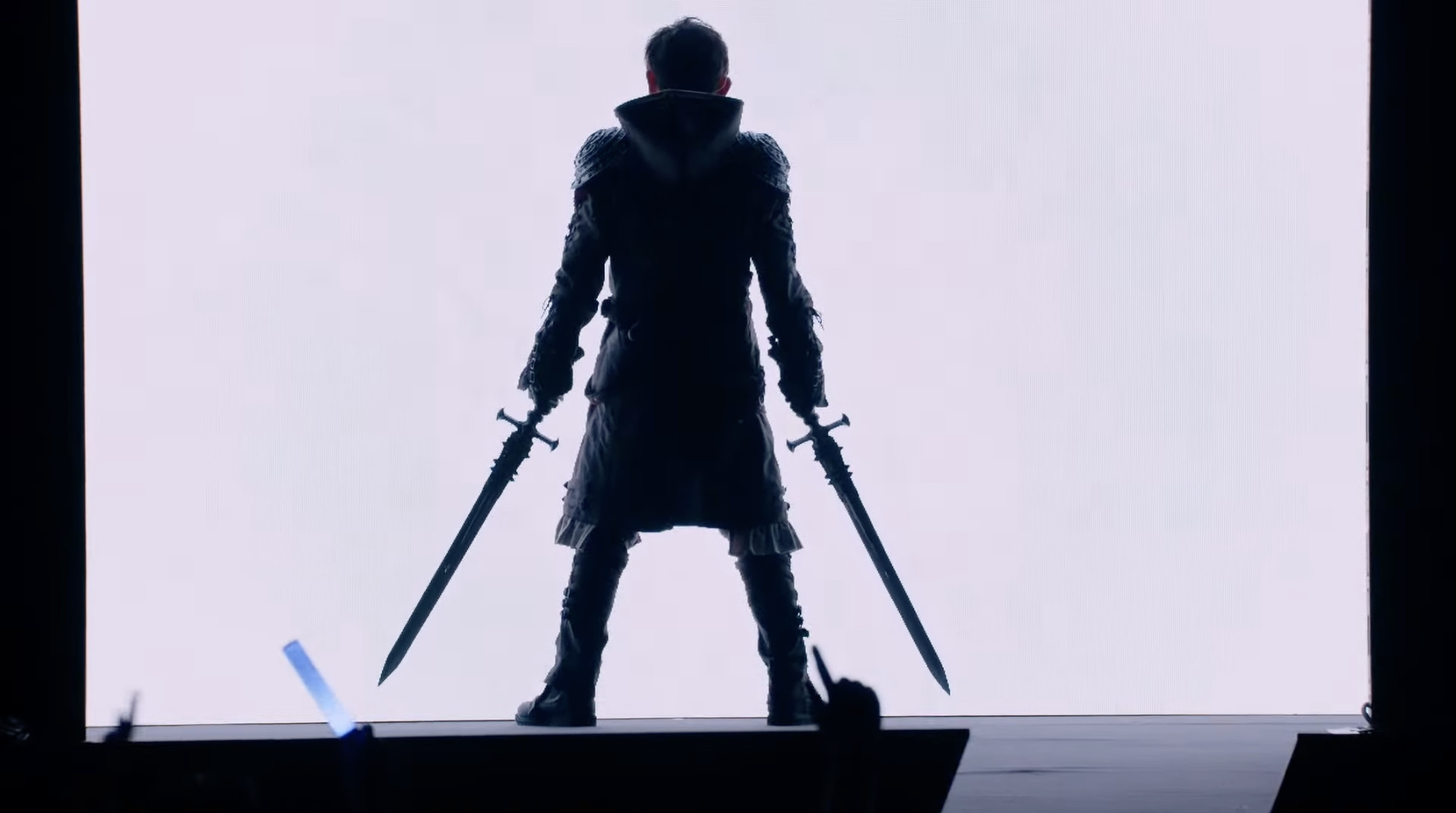 Image from the 2023 Final Fantasy XIV Fan Festival London featuring game producer Naoki Yoshida standing silhouetted against a white screen holding two swords dressed as the new Viper DPS class from FFXIV.